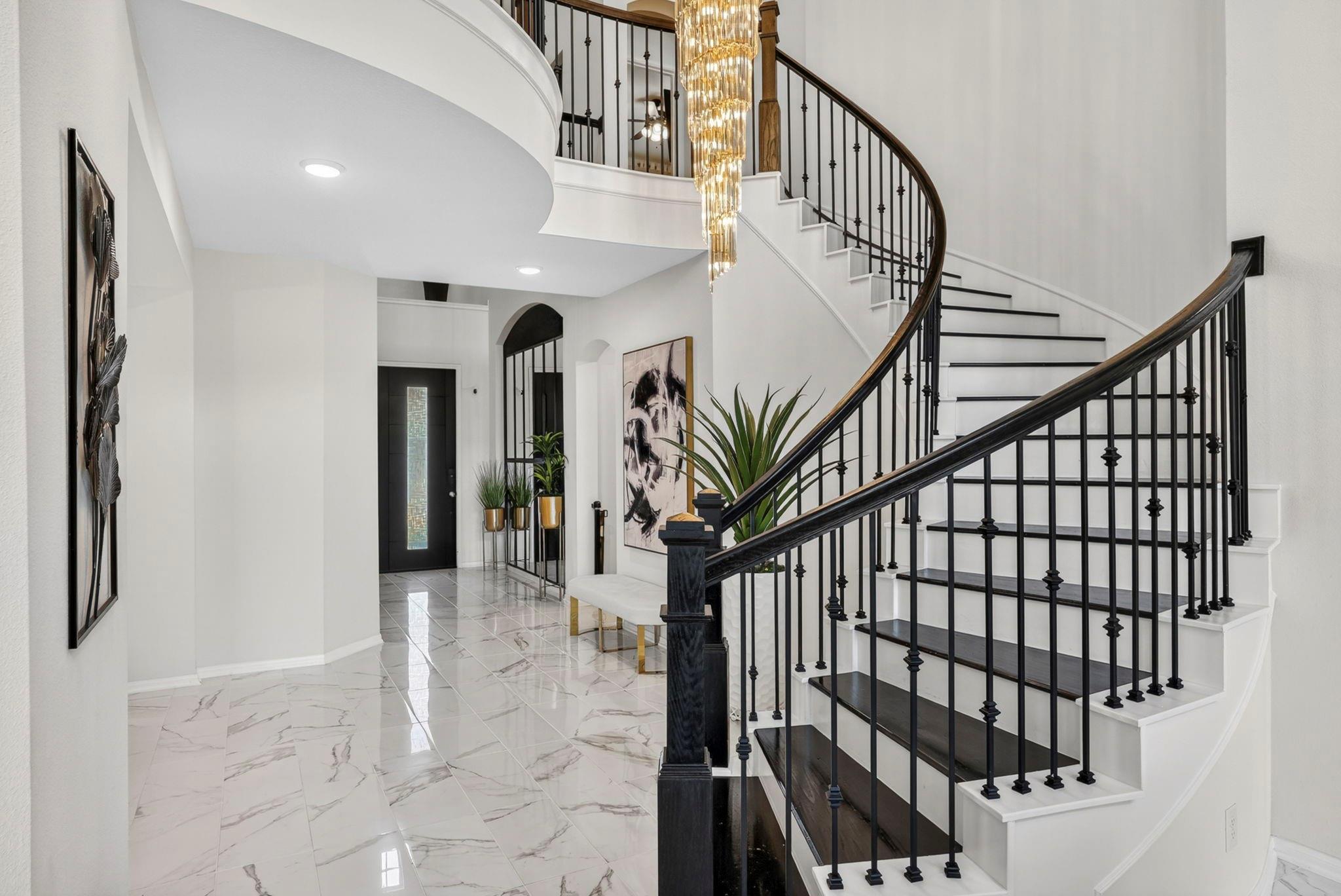 Entry foyer with a curved staircase, black iron railings, glossy tile floors, and a cascading chandelier.