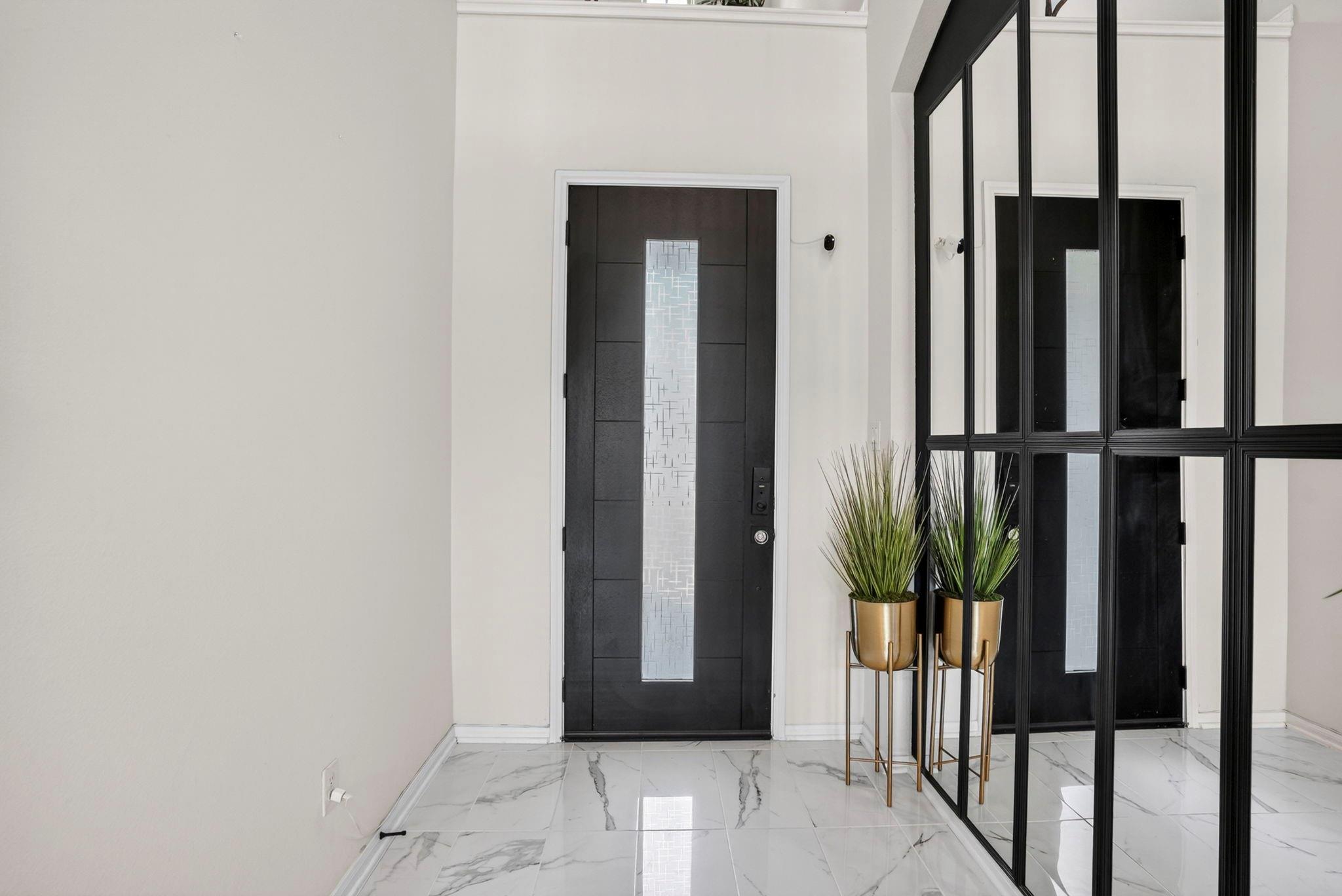 Entry foyer with a black glass-panel door, marble-look tile, mirrored partition, and tall potted plants.