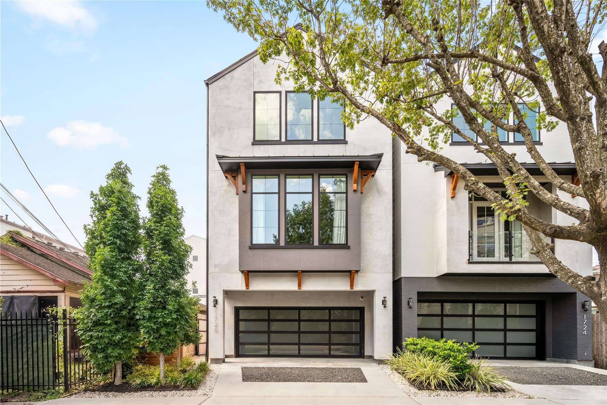 Modern three-story home with white and gray facade, black-framed windows, and a wide garage beneath a leafy tree.