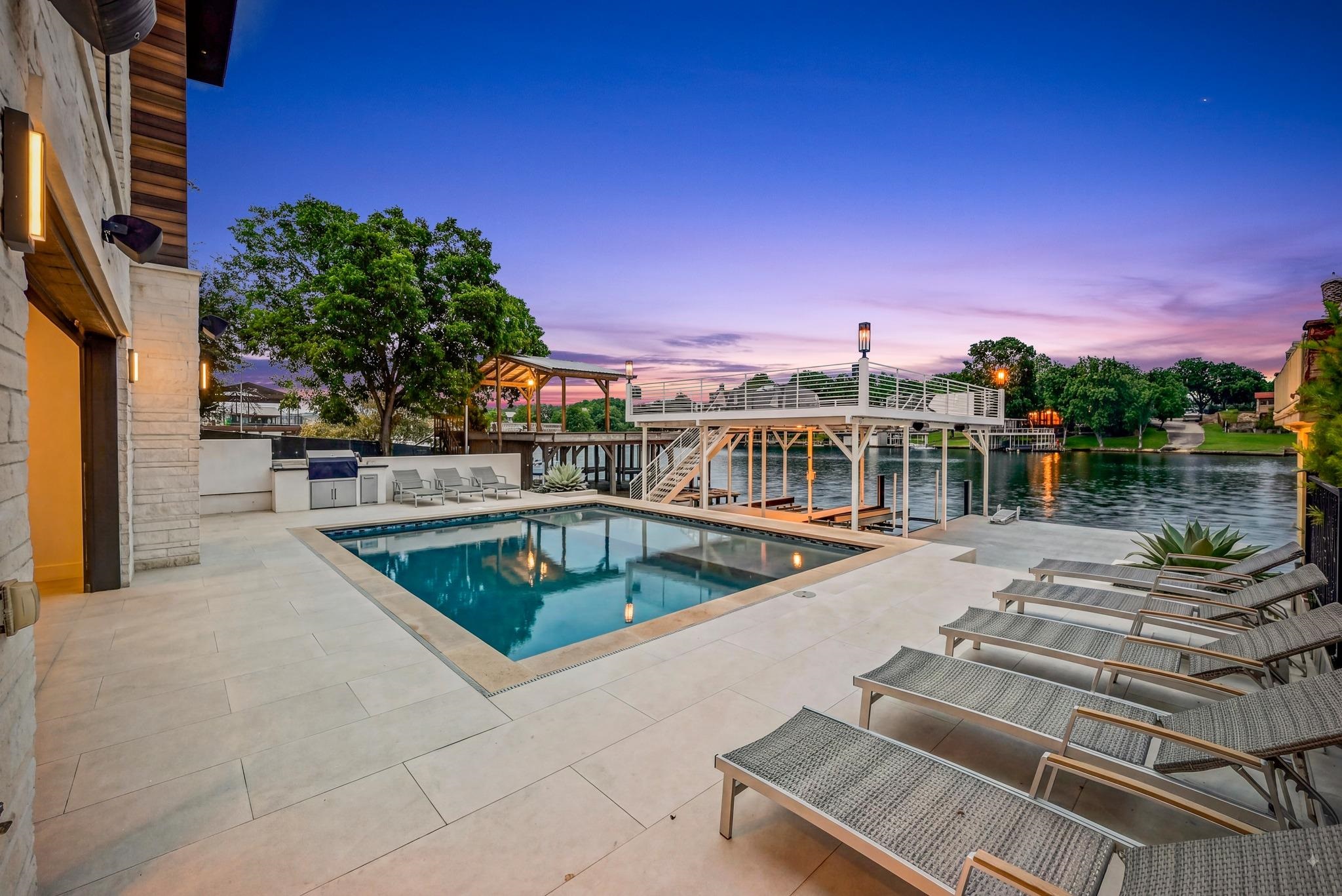 Lakeside patio with a swimming pool, lounge chairs, and raised dock at dusk.