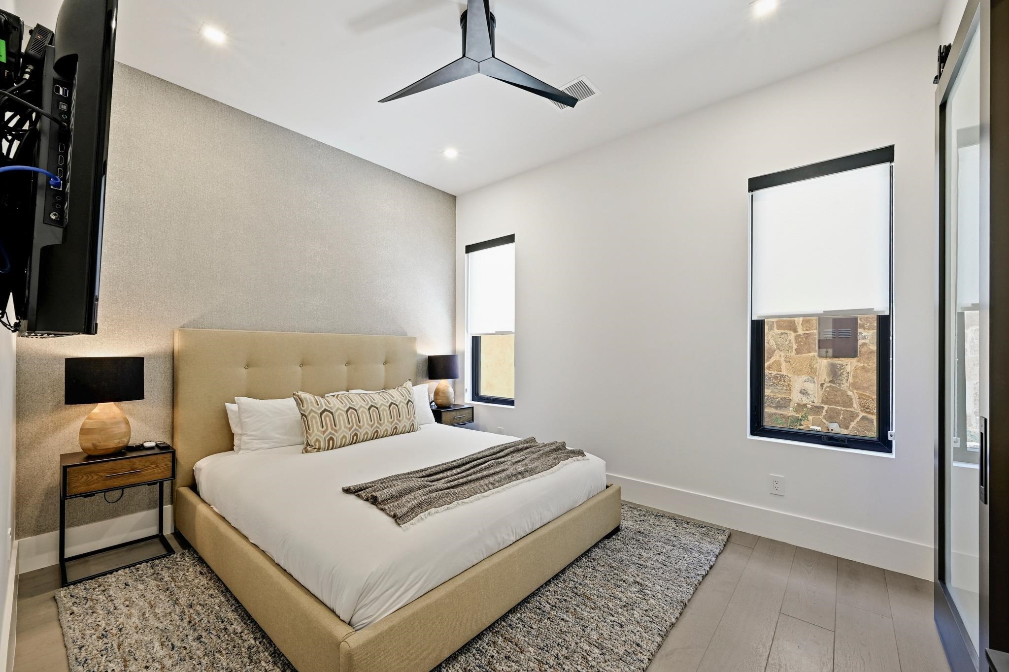 Staged bedroom with a beige upholstered bed, patterned pillows, gray rugs, and shaded windows.