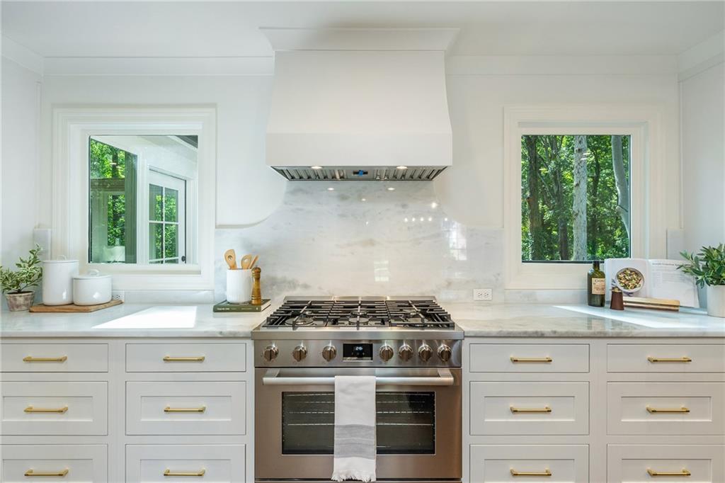 White kitchen with a stainless steel range, marble backsplash, brass cabinet pulls, and tree views through two windows.