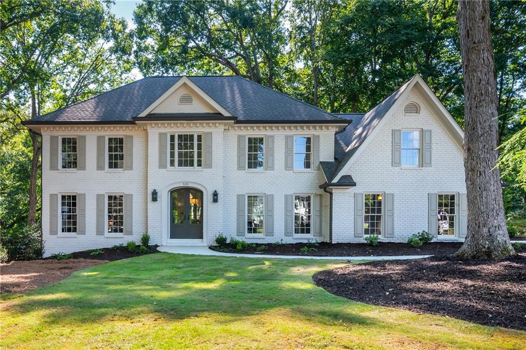 White two-story brick house with gray shutters, a dark roof, and a tree-shaded lawn.