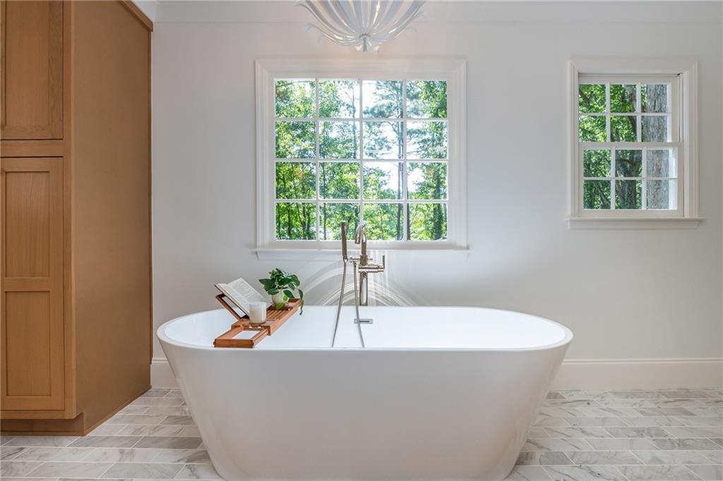 Freestanding soaking tub beneath two windows, with warm wood cabinetry and marble floor tile.