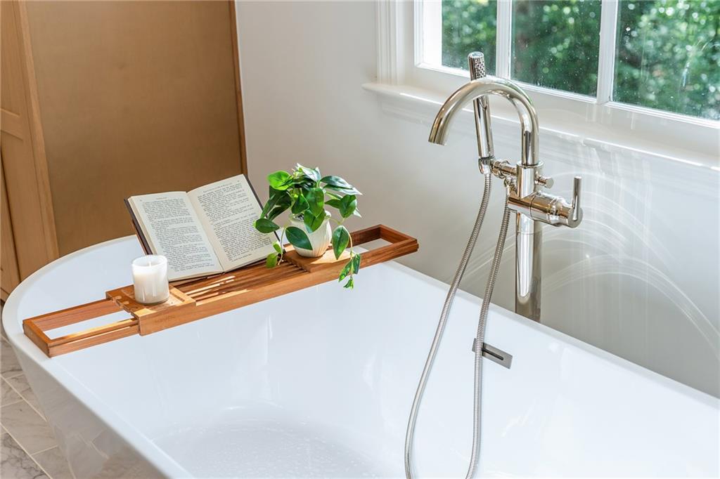 Soaking tub beneath a large window, with a chrome shower fixture and a wooden tray holding a book and plant.