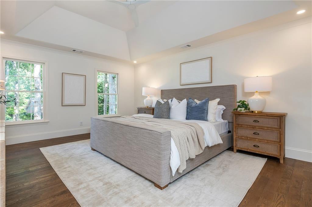 Staged bedroom with an upholstered bed, blue accent pillows, wooden dresser, and tall windows overlooking trees.
