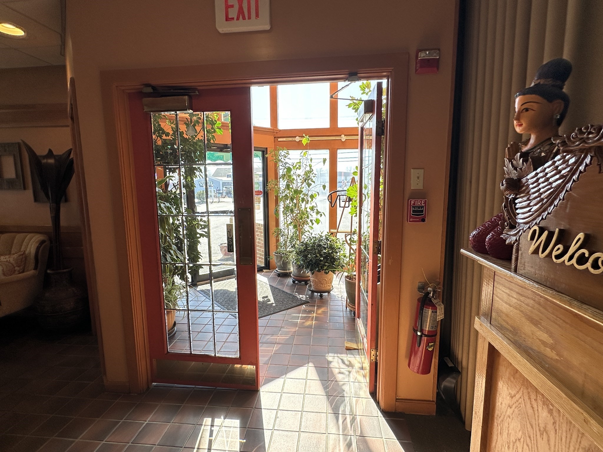 Commercial entryway with a red-framed glass door opening into a bright, plant-filled tiled sunroom.