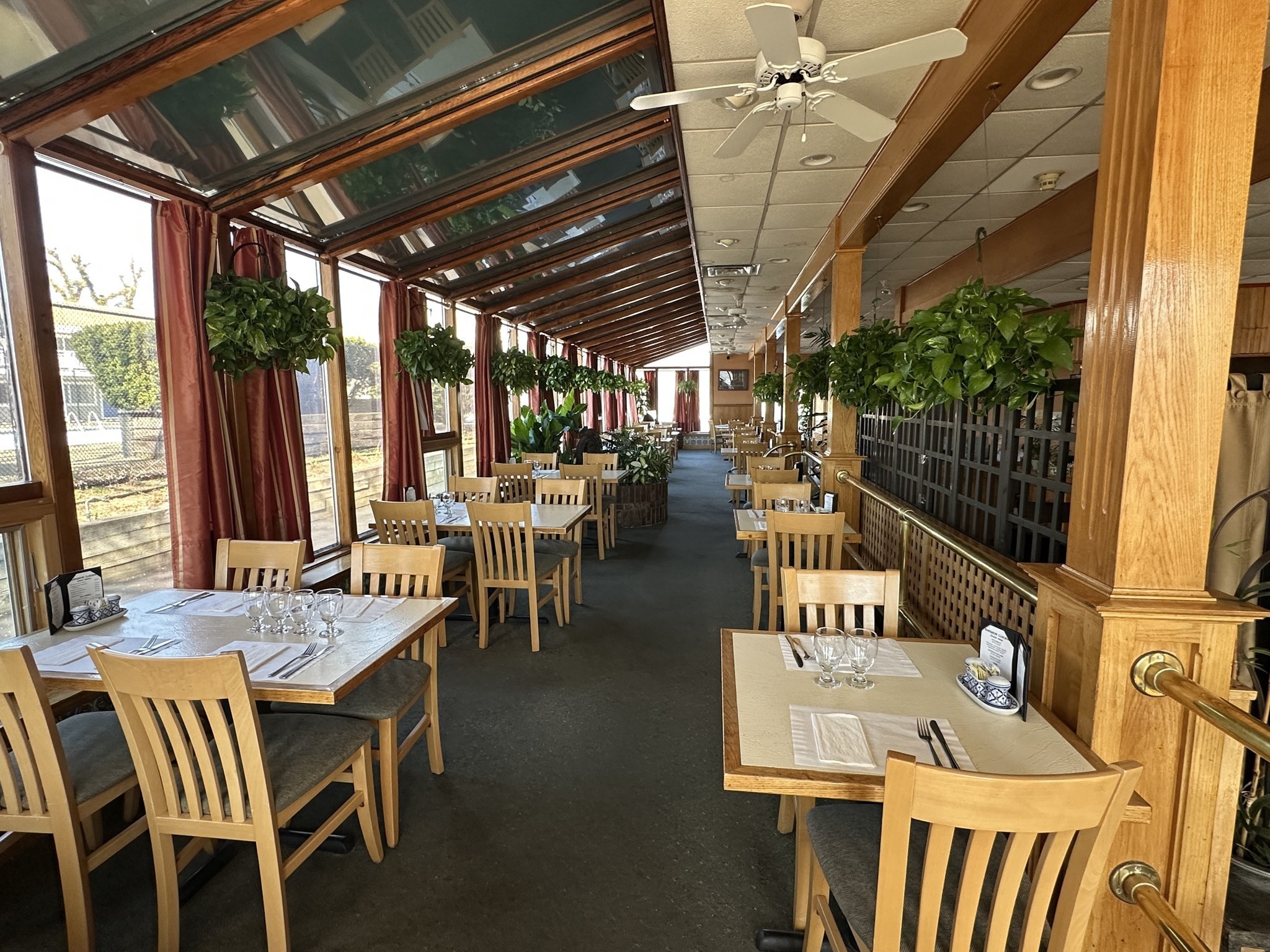 Restaurant dining room with set tables beneath a sloped glass roof and rows of potted plants.