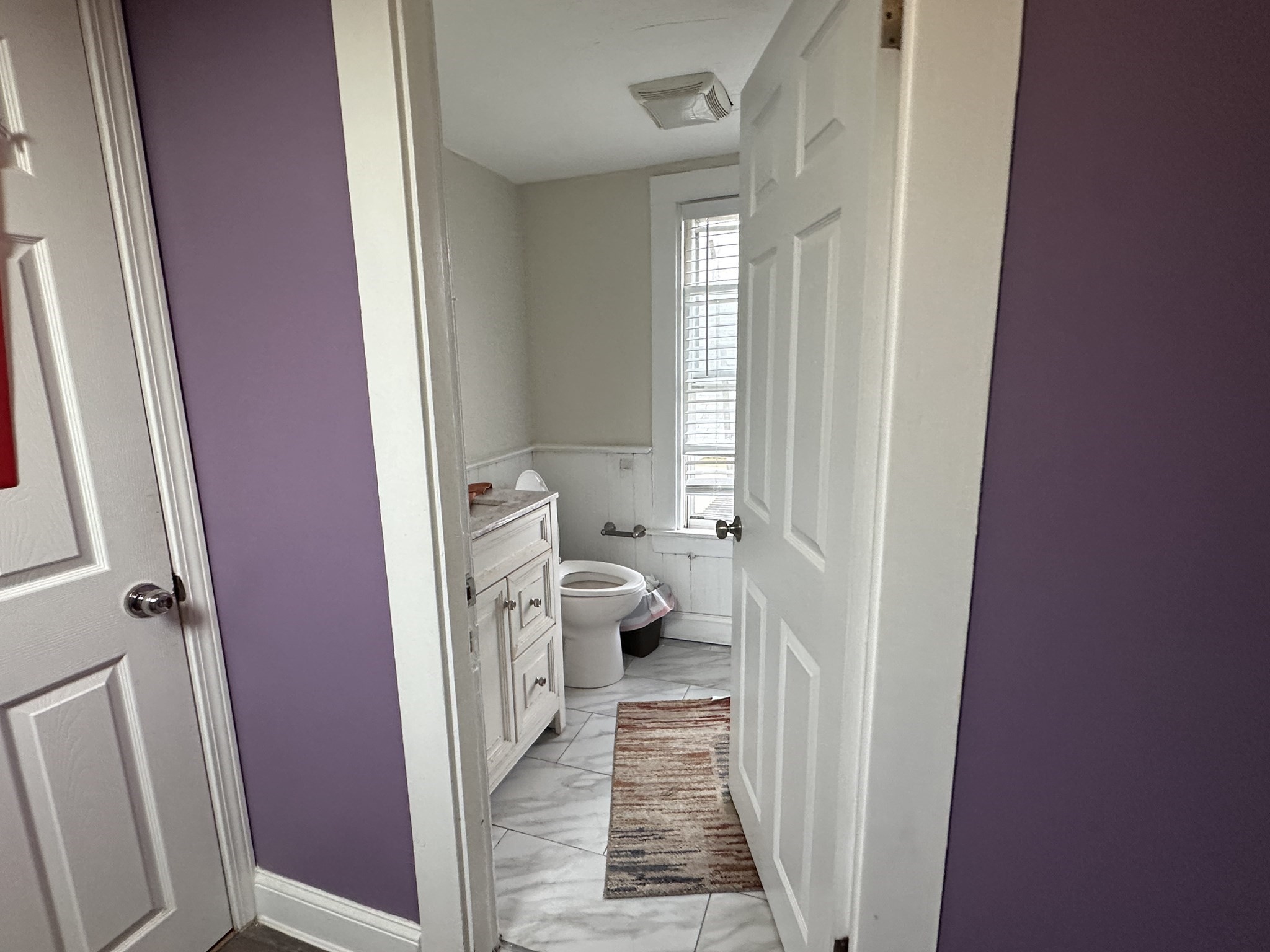 Bathroom with a white vanity, toilet, narrow window, and striped runner.