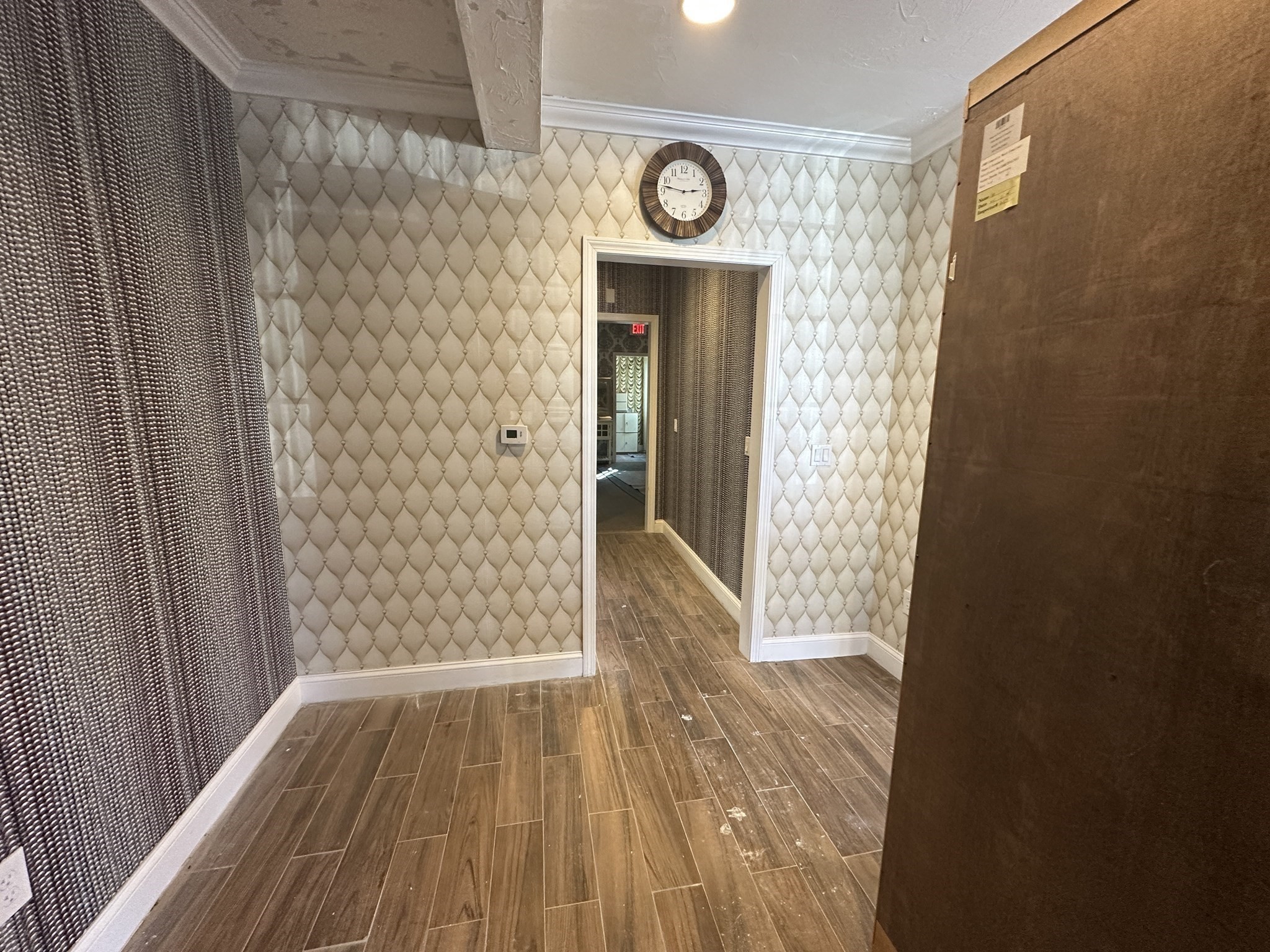 Hallway with quilted wallpaper, brown tile flooring, and a doorway beneath a round clock.