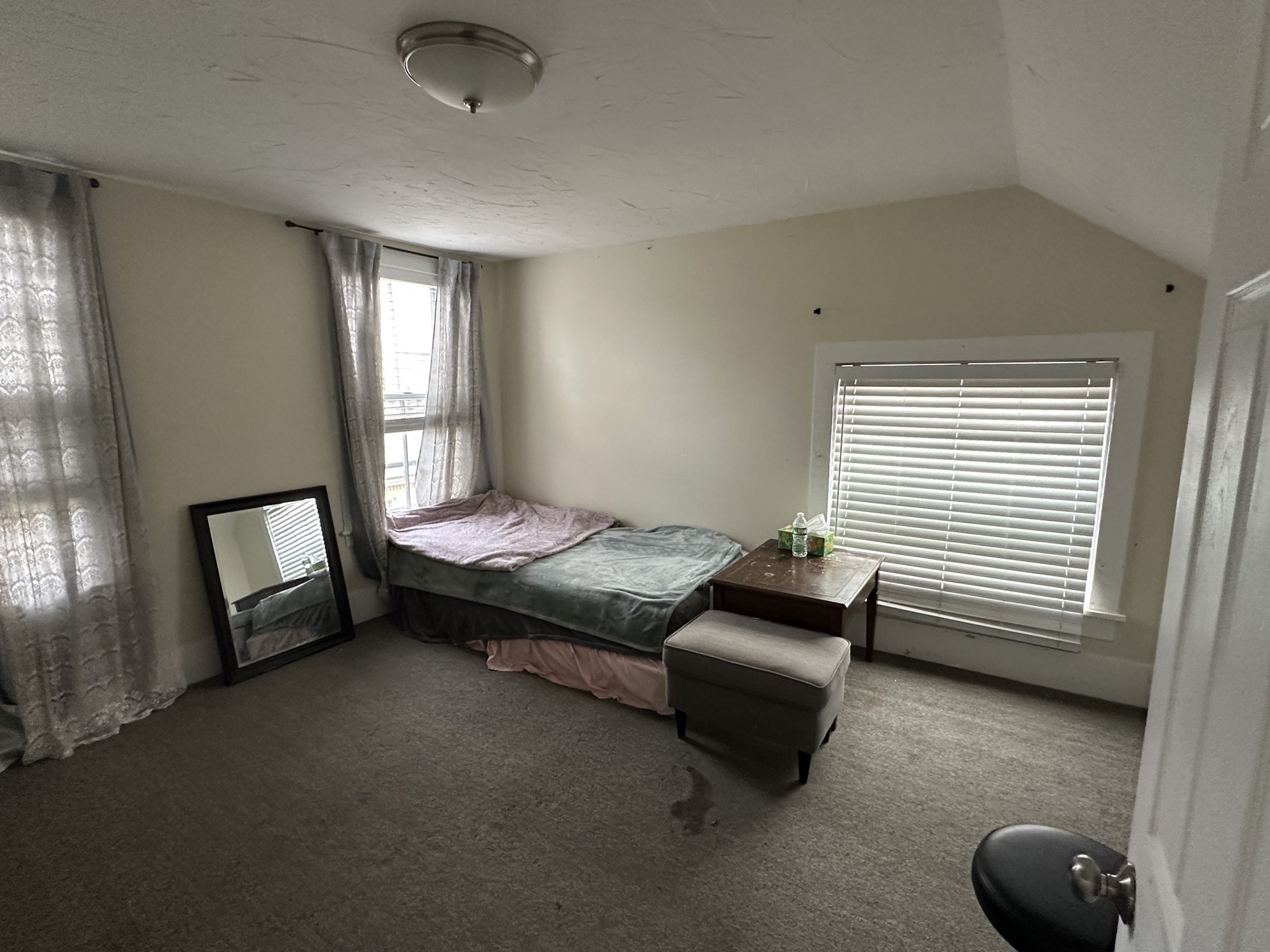 Bedroom with sloped ceiling, single bed, carpet, and two windows with curtains and blinds.