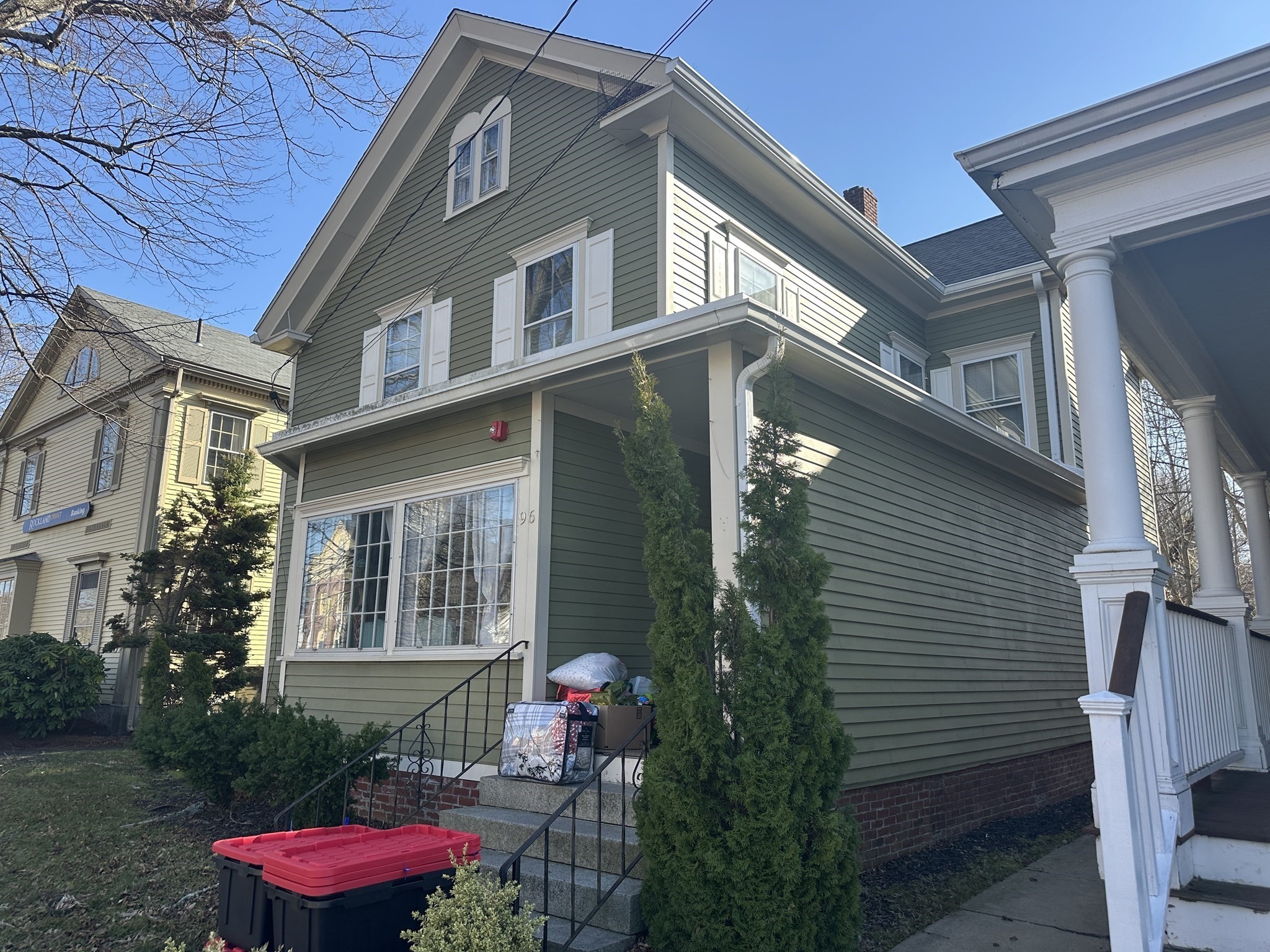 Green two-story house with white trim, steep gable, and evergreen shrubs beside the front steps.