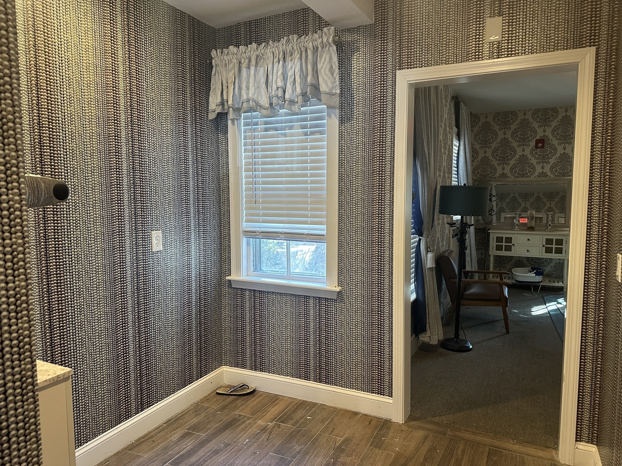 Bathroom with textured wallpaper, wood-look tile flooring, and a window overlooking a furnished adjoining room.