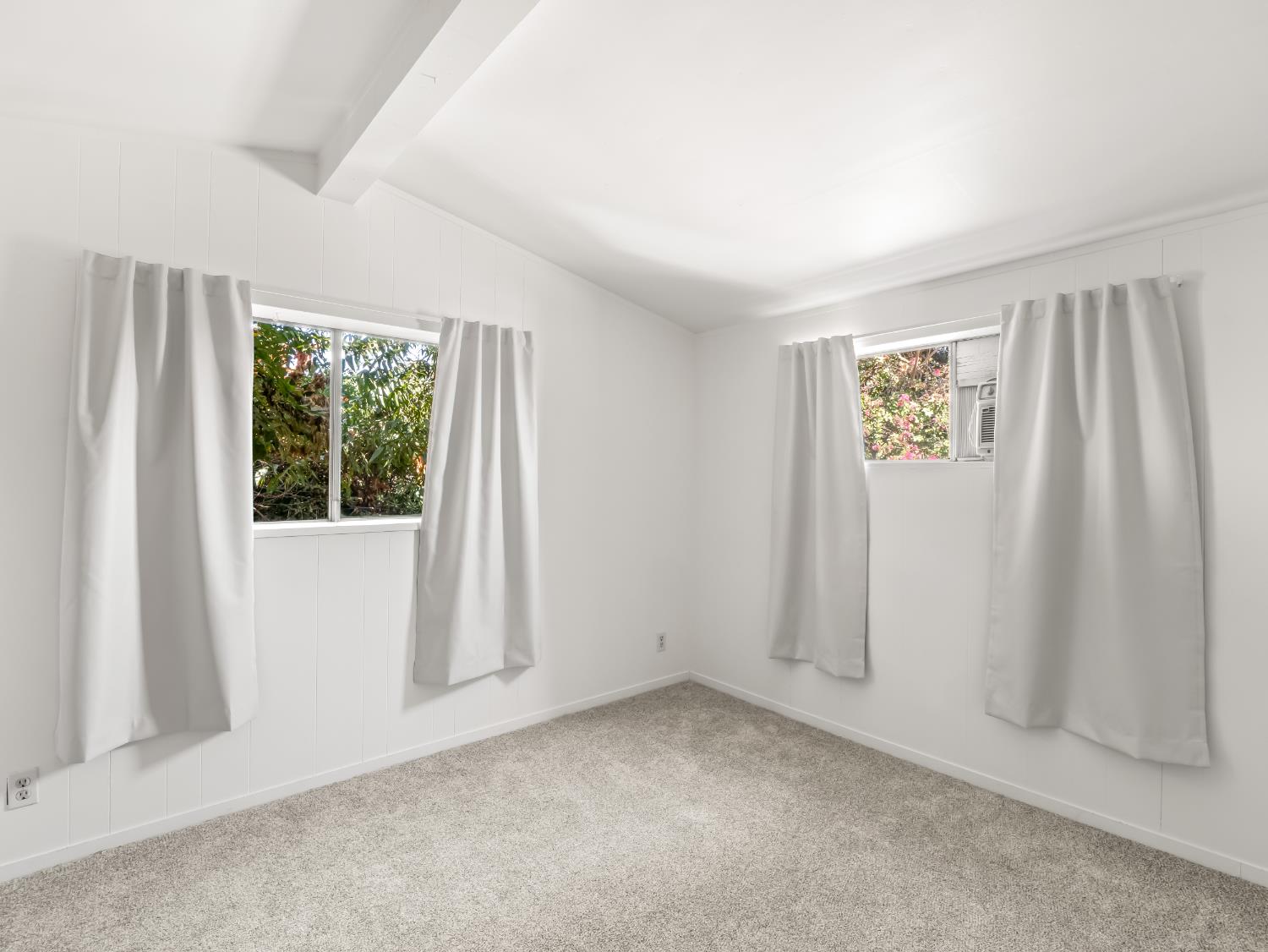 Empty bedroom with gray carpet, white curtains, and two windows overlooking greenery.