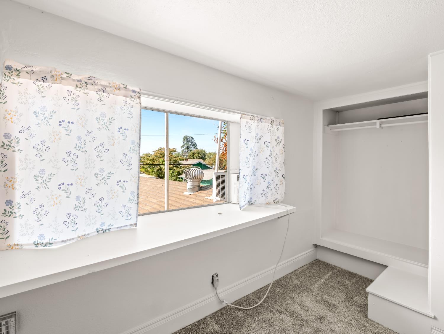 Empty carpeted bedroom with floral curtains, a wide window, and an open closet.