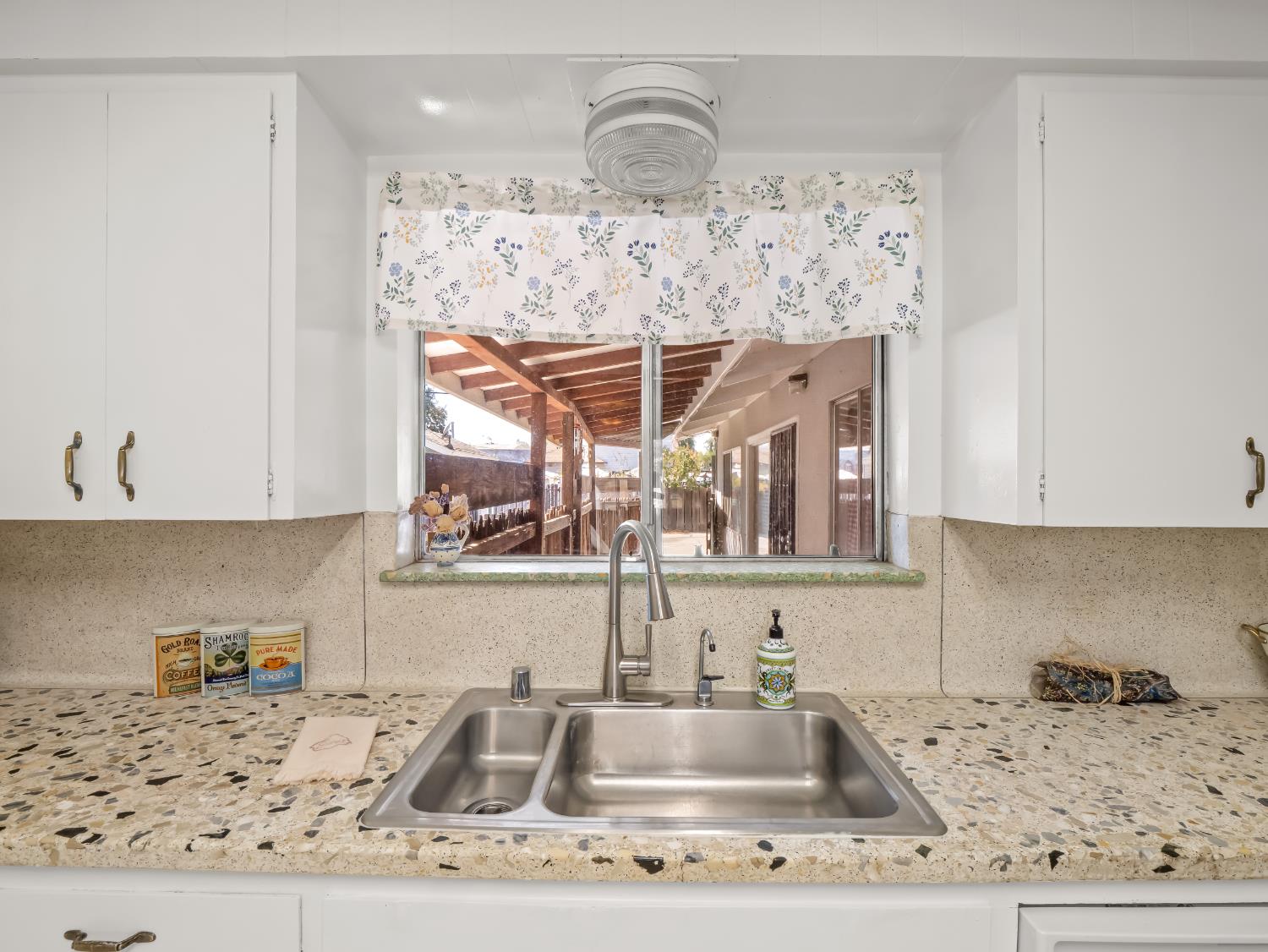 Kitchen sink beneath a window overlooking a covered patio, with white cabinets and a speckled countertop.