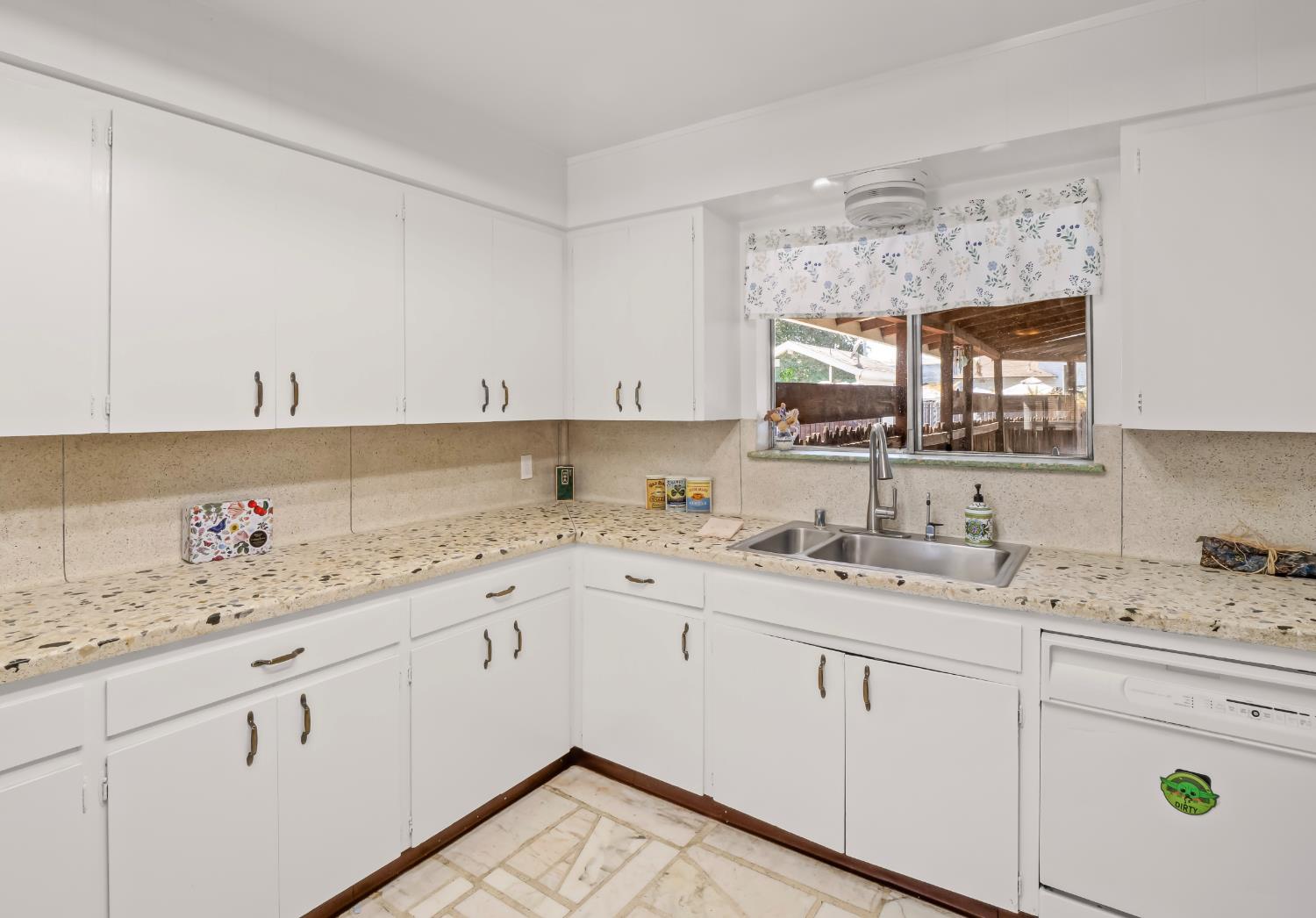 Kitchen with white cabinets, speckled countertops, a double sink, and a window overlooking a covered patio.