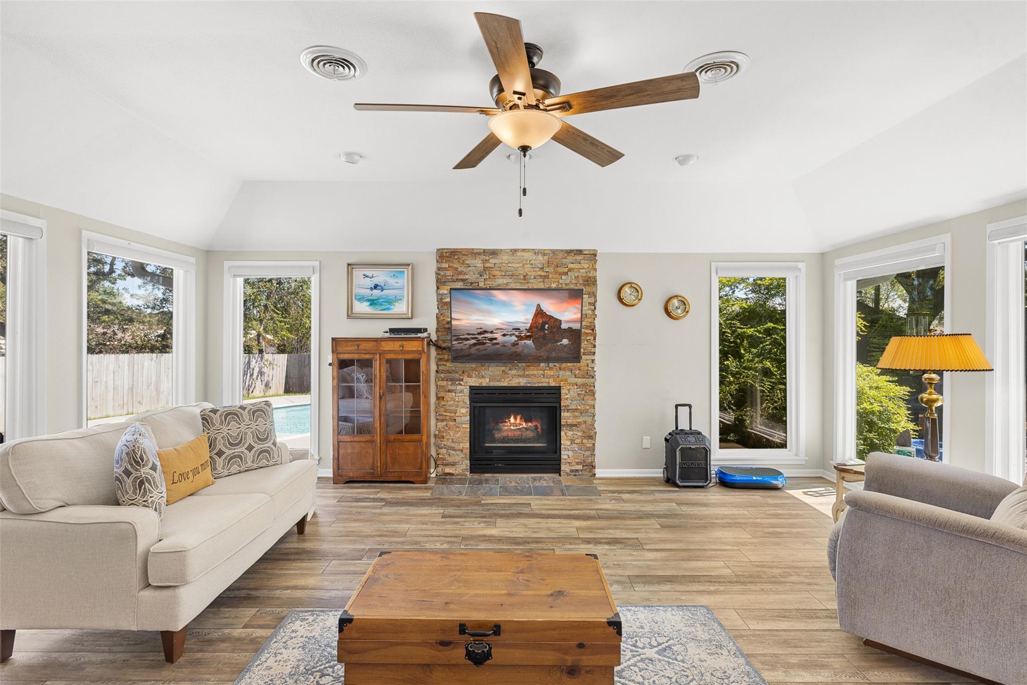 Living room with a stone fireplace, vaulted ceiling, ceiling fan, and windows overlooking a pool.