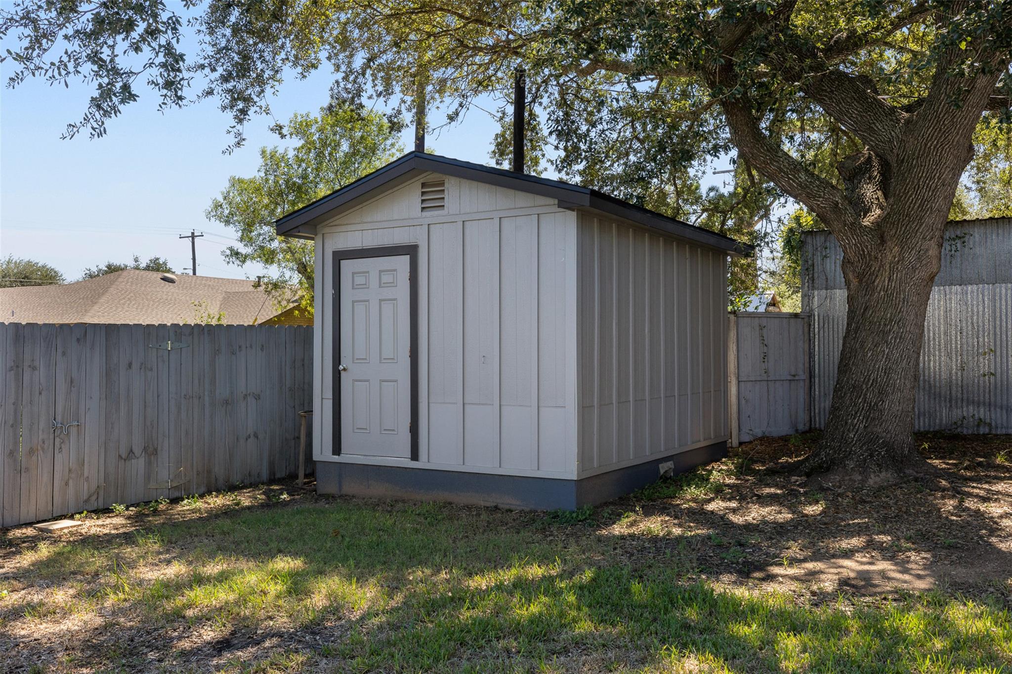 Detached light-gray storage shed with a dark-trimmed gabled roof beneath a large oak tree.
