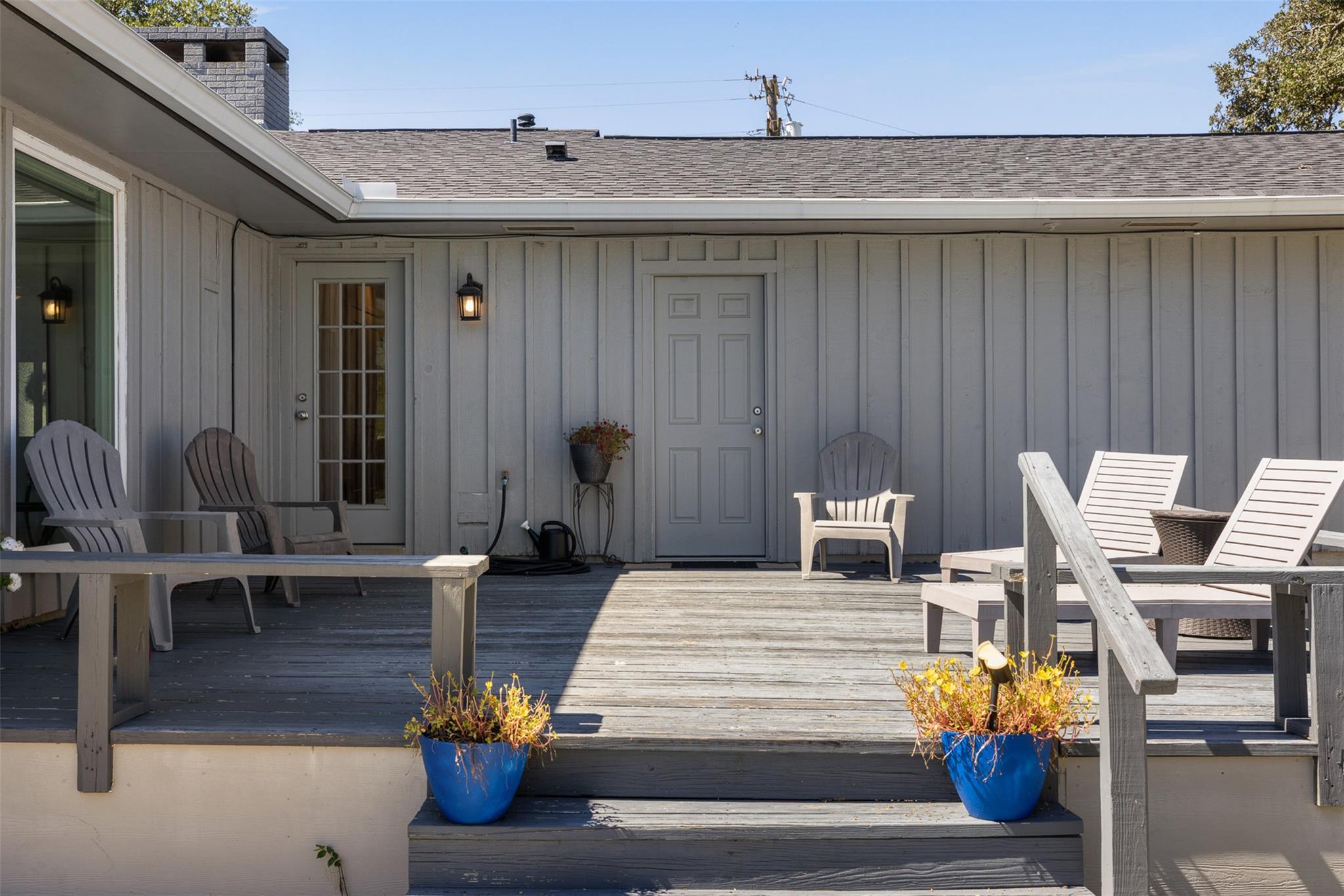 Furnished wooden deck with gray siding, Adirondack chairs, blue planters, and steps to the patio.