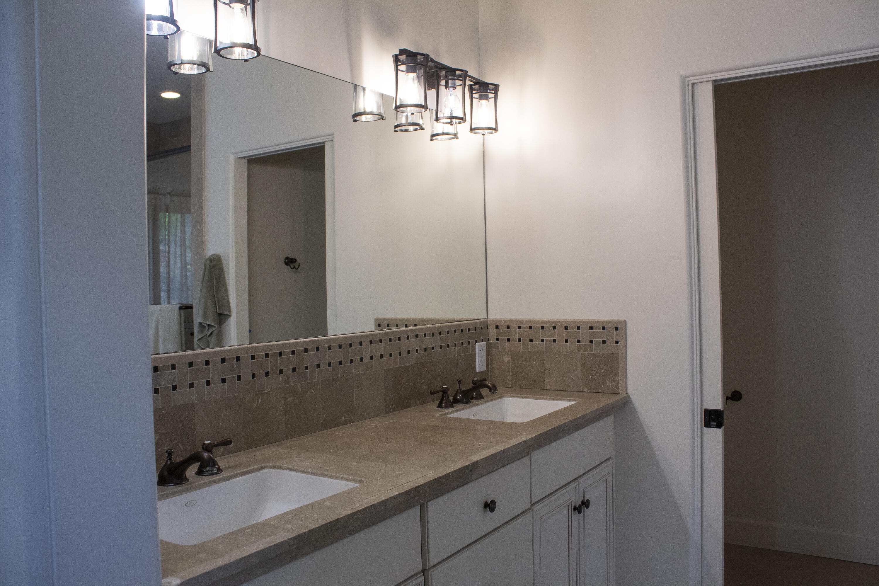 Bathroom vanity with double sinks, taupe stone counters, mosaic tile trim, and pendant lights.