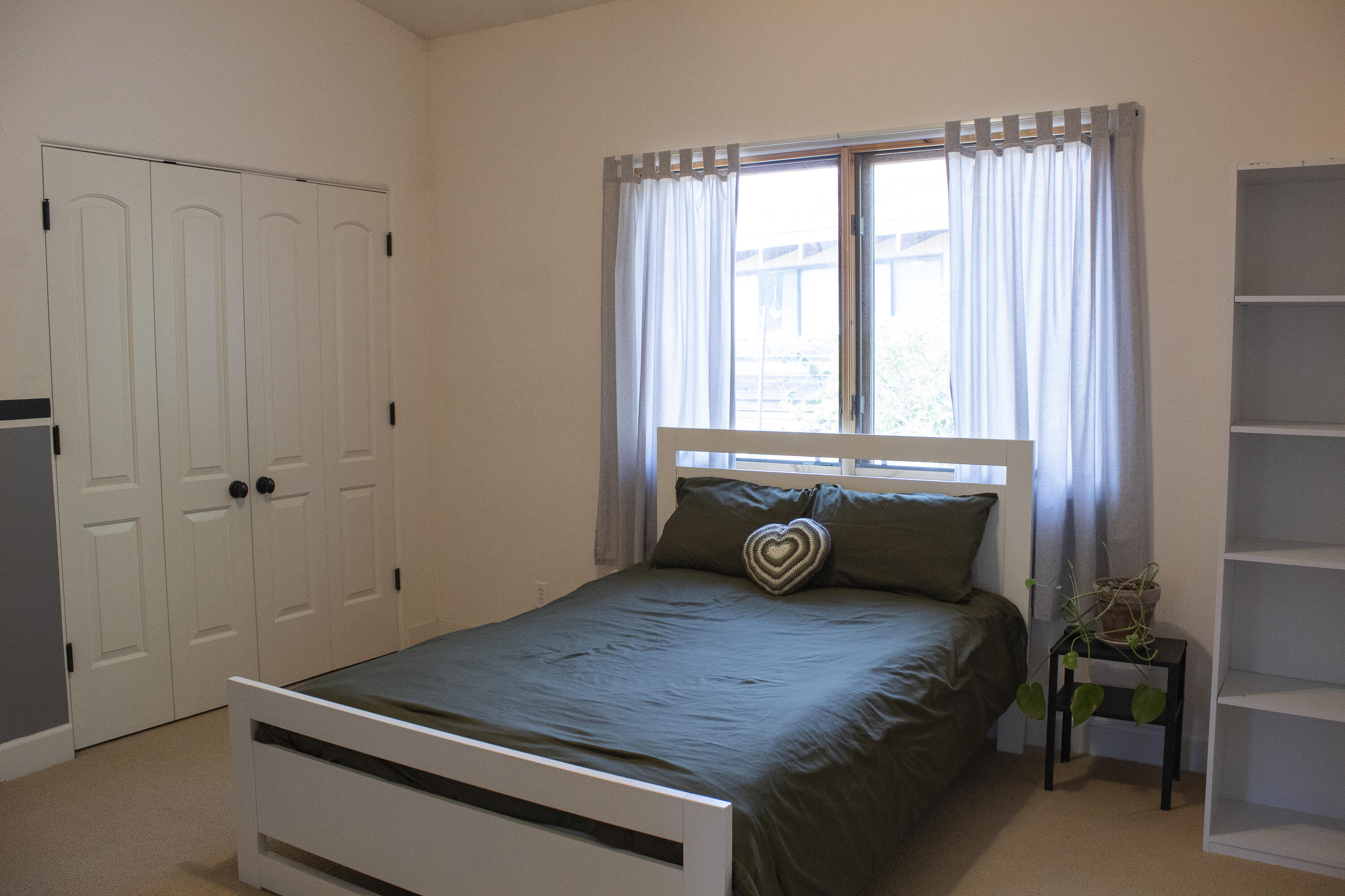 Bedroom with a white bed, dark bedding, closet doors, and a curtained window.