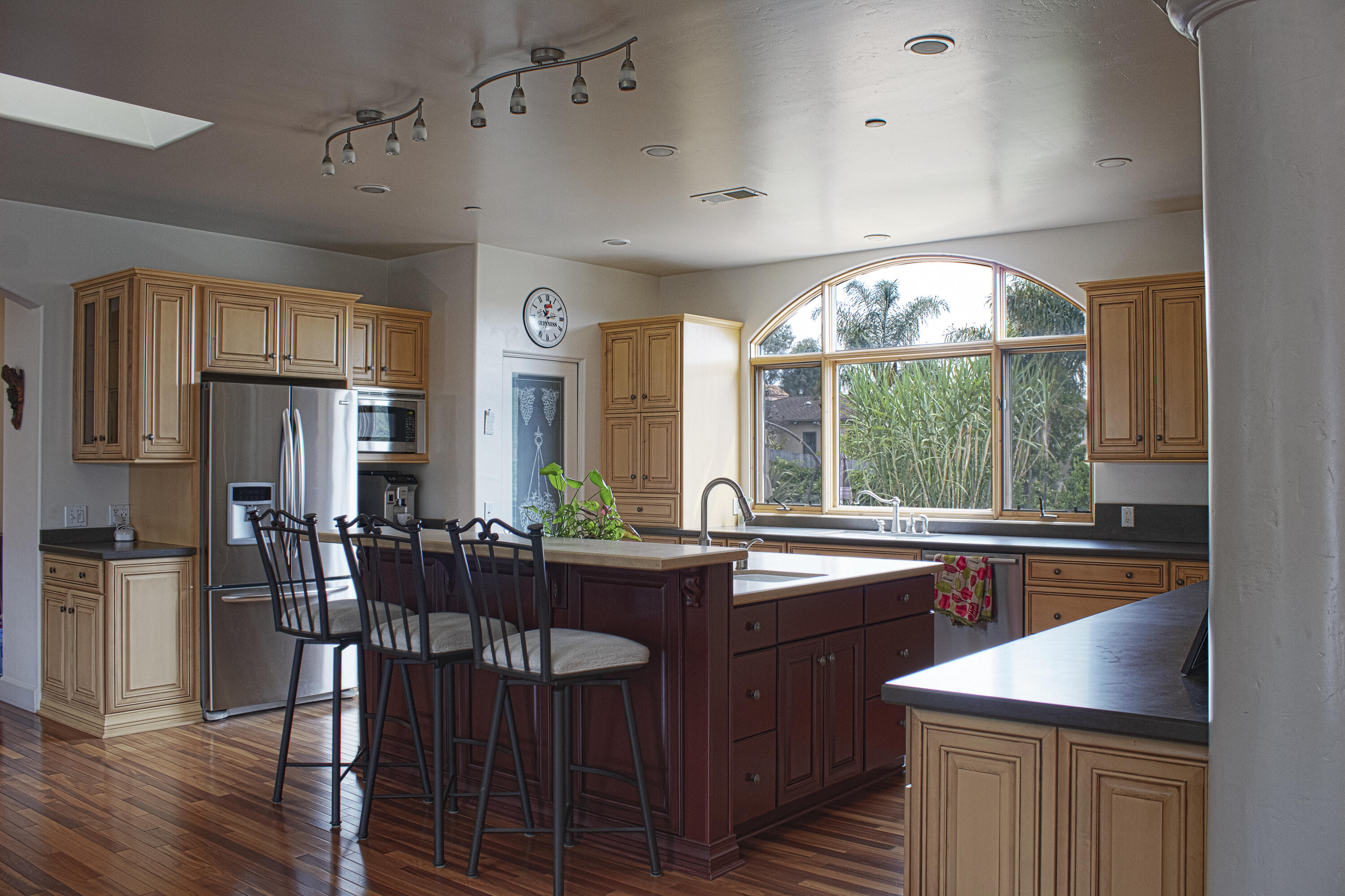 Kitchen with a dark red island, bar stools, stainless appliances, and a large arched window.