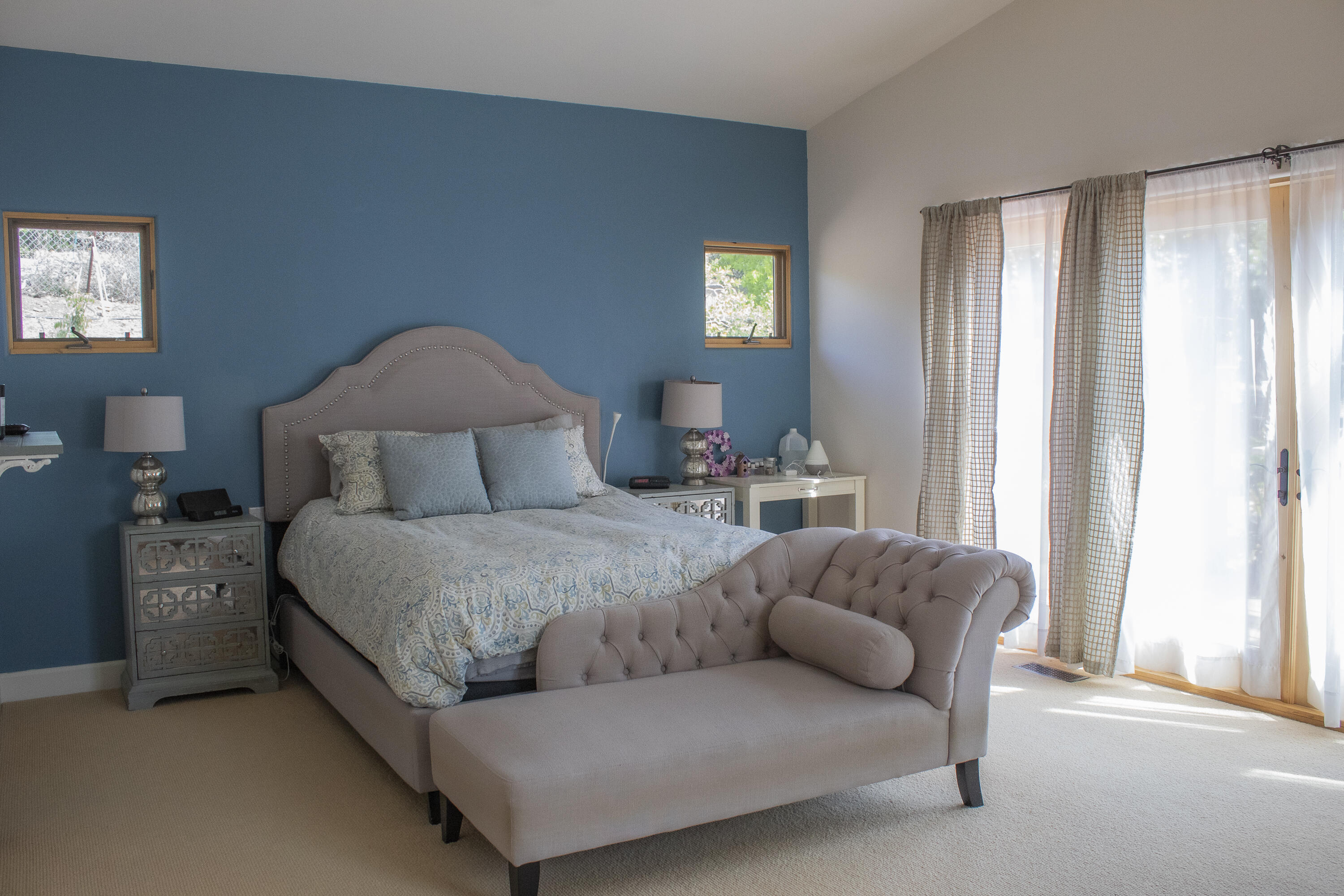 Bedroom with a blue accent wall, patterned bedding, tufted chaise, and bright curtained glass doors.