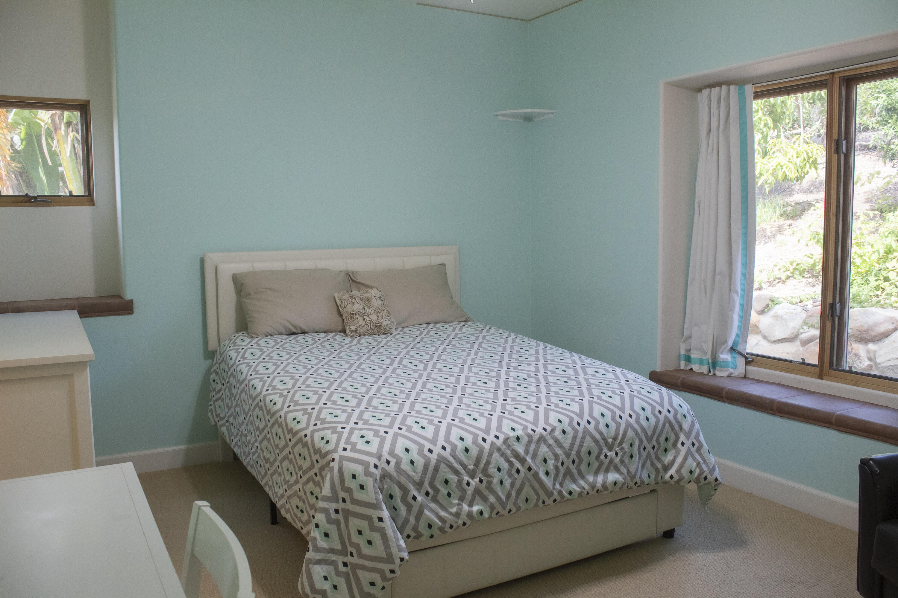 Bedroom with turquoise walls, geometric bedding, and a cushioned window seat beside large windows.