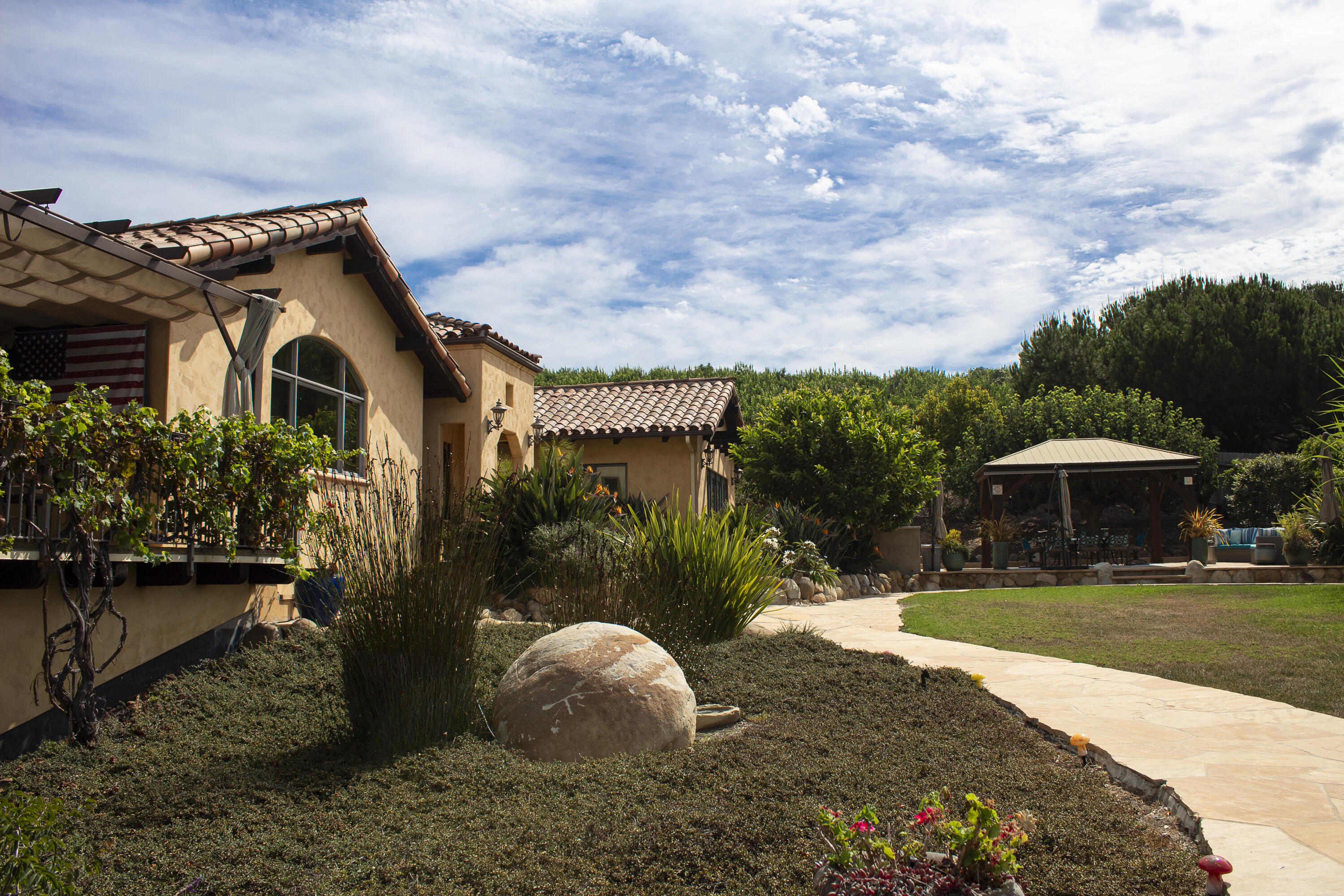 Tan stucco home with a vine-covered patio, landscaped yard, and gazebo beside a curved stone walkway.