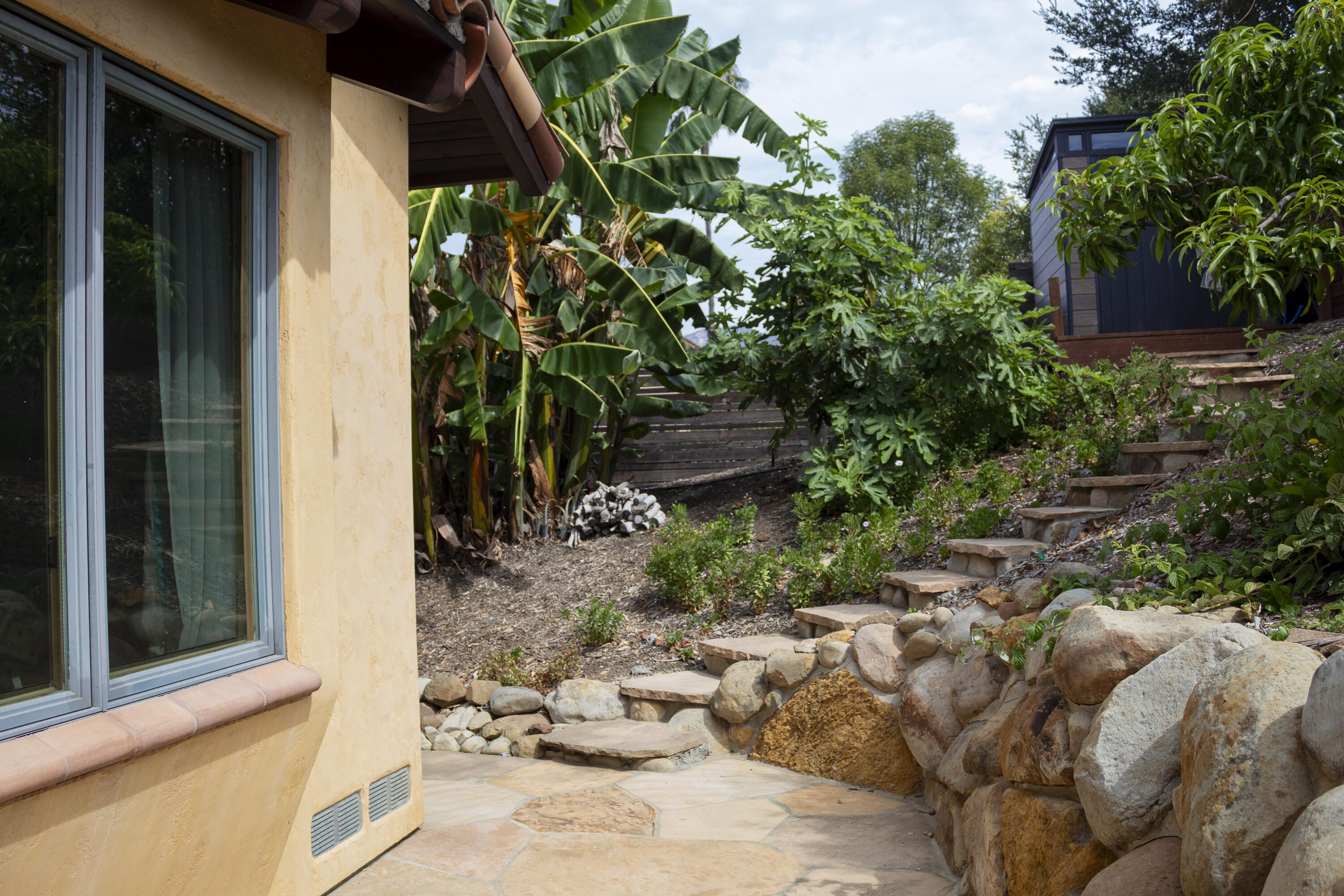 Side yard with flagstone patio, stone steps, boulder retaining wall, and lush tropical plants.