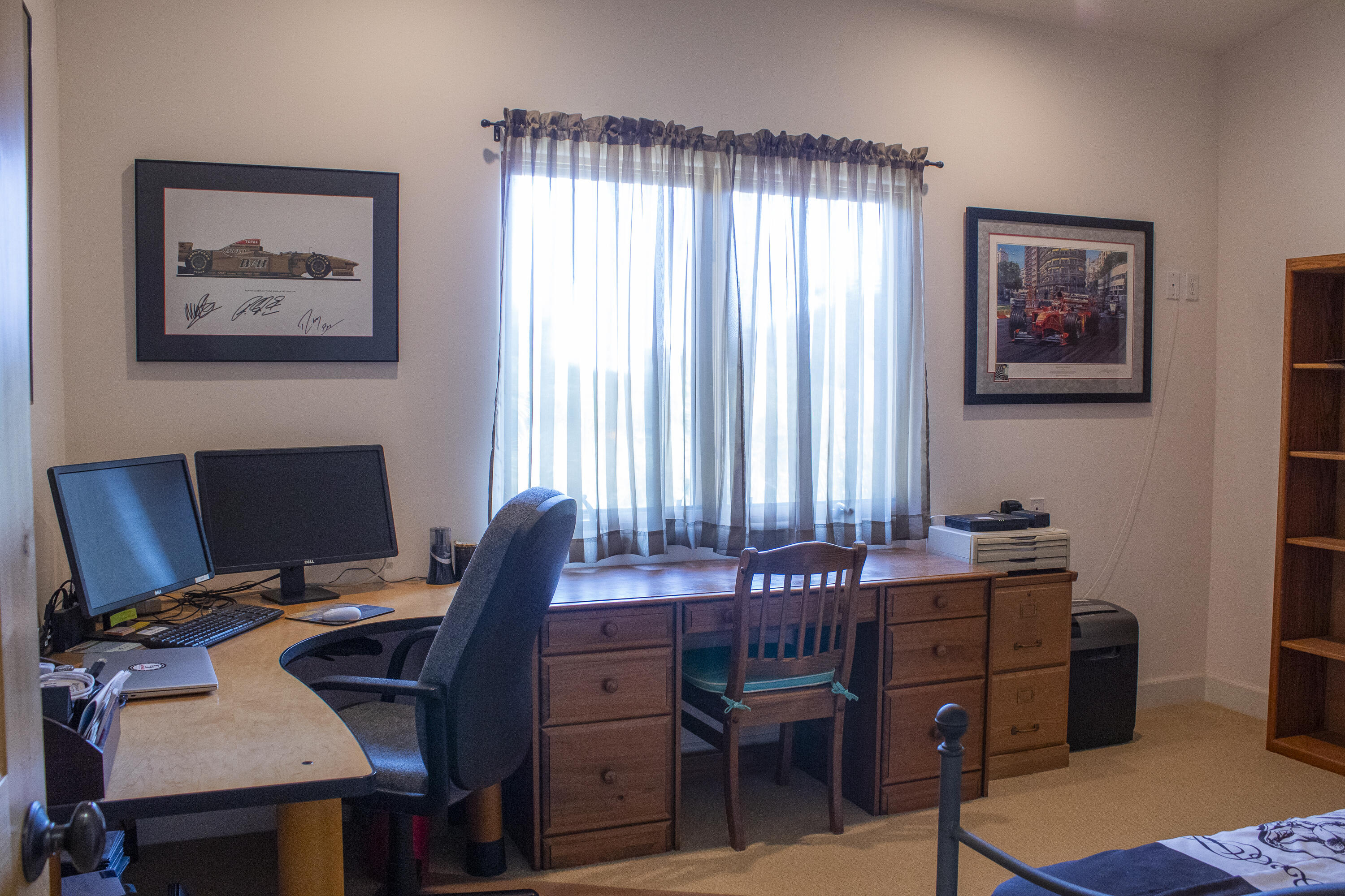 Furnished home office with a long wooden desk, dual monitors, and a curtained window.