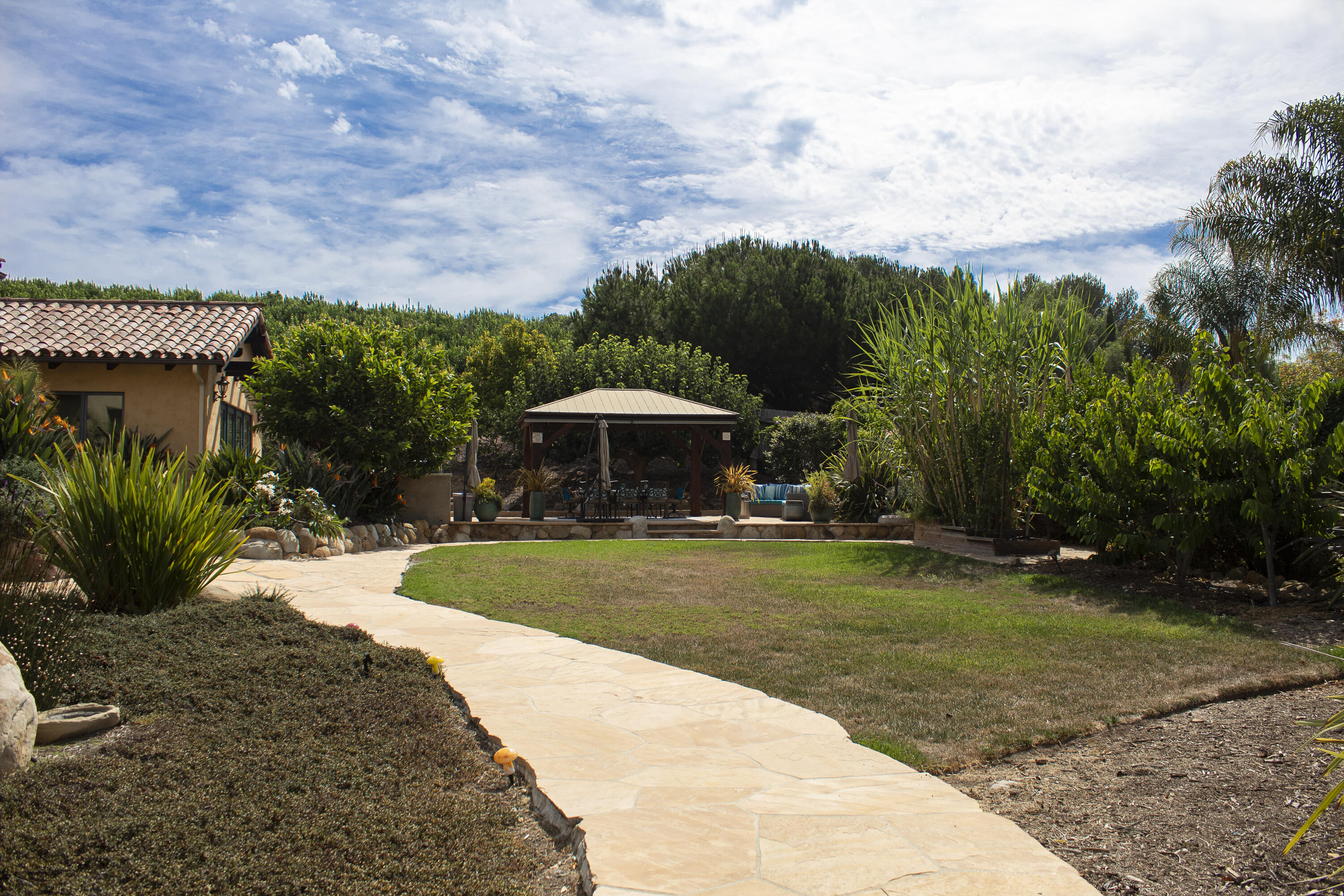 Landscaped backyard with a curving stone patio, lawn, and covered pergola surrounded by lush greenery.