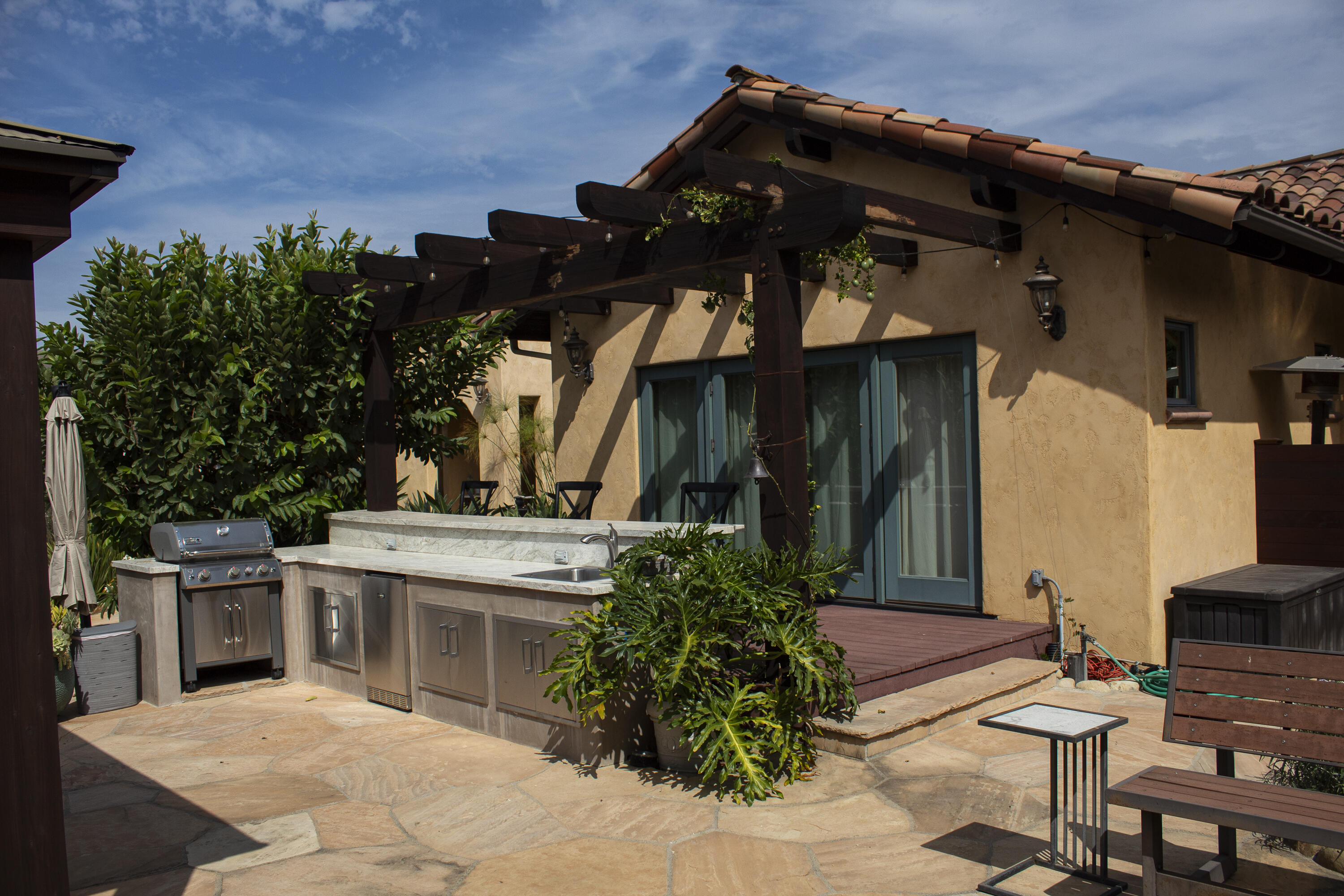 Outdoor kitchen with stainless steel grill beneath a pergola beside the home’s stone patio.