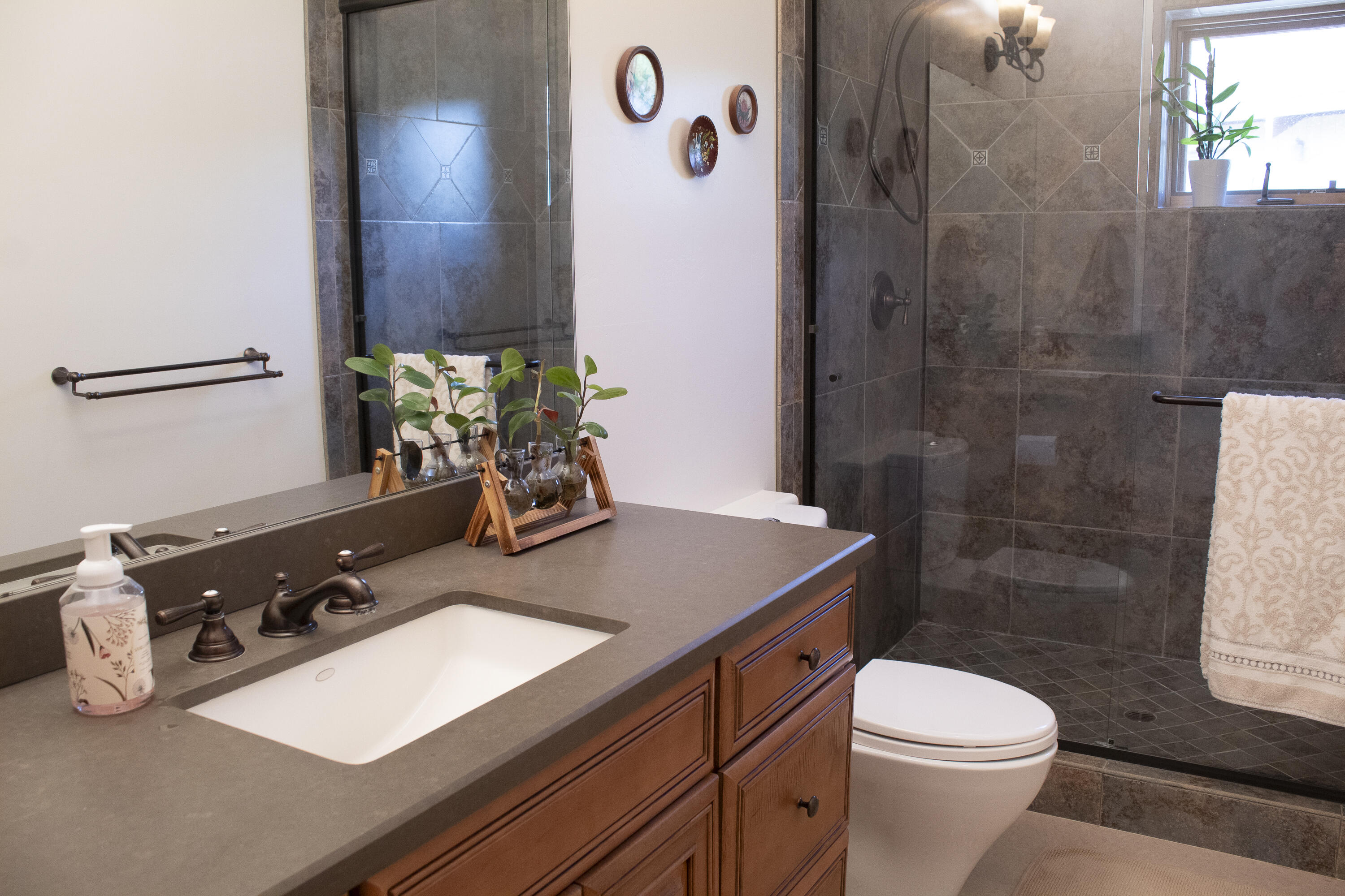 Bathroom with a wood vanity, gray countertop, dark tiled walk-in shower, and small window.