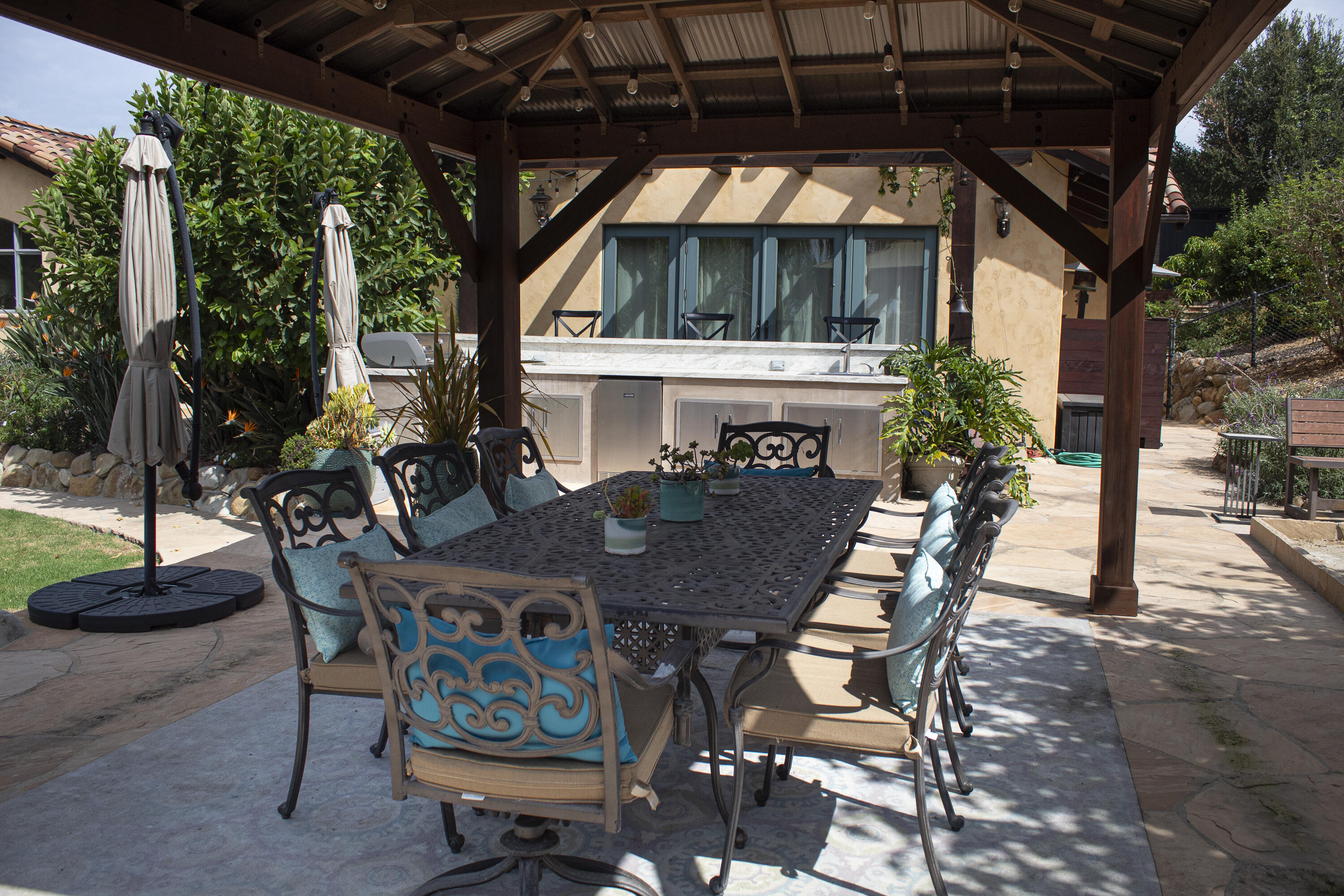 Outdoor dining patio beneath a timber pergola, with a long table and outdoor kitchen counter.