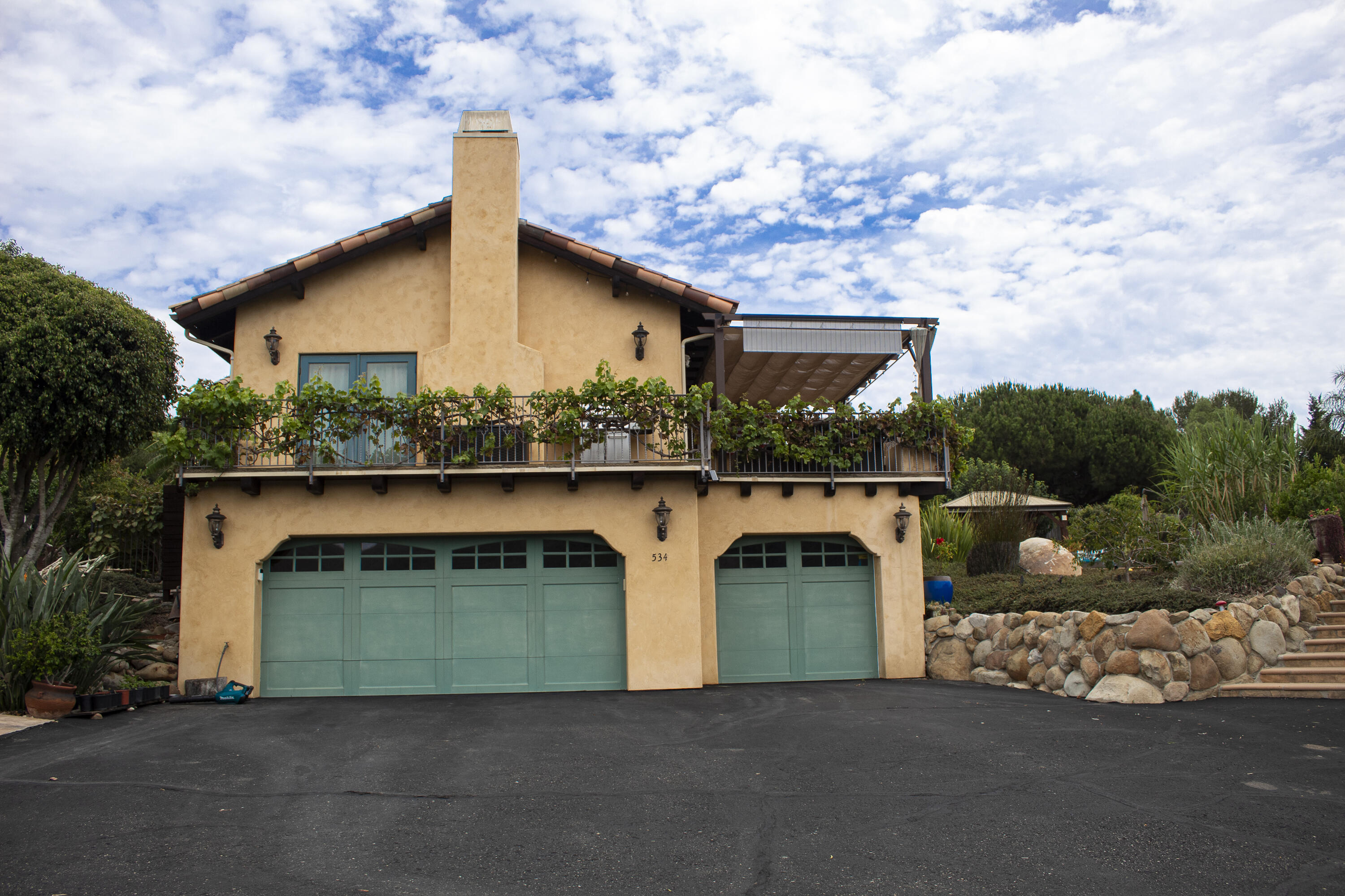 Tan stucco home with teal garage doors, a vine-covered balcony, and a stone garden wall.