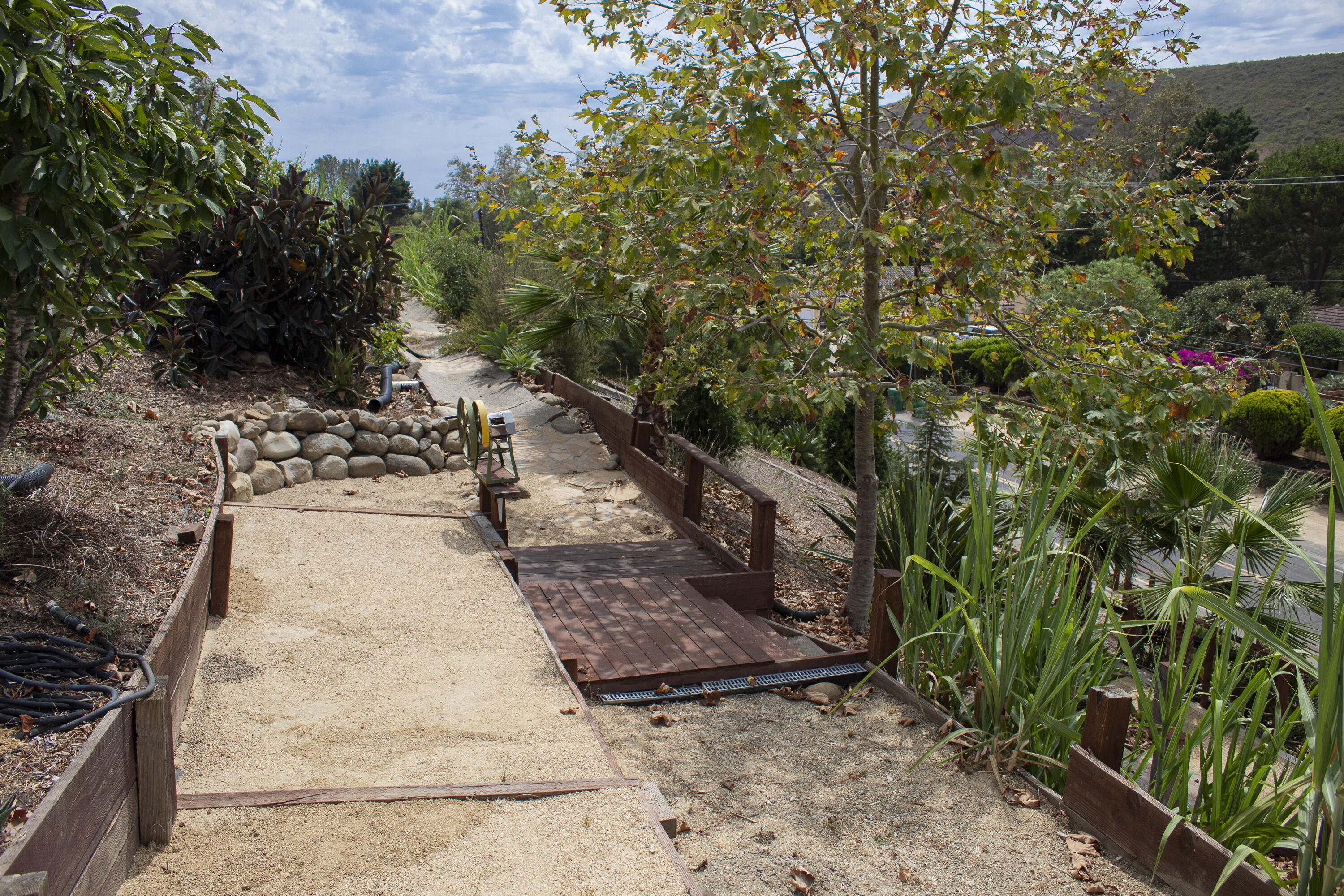 Sloping landscaped yard with a gravel path, wooden steps, and mature trees and plants.