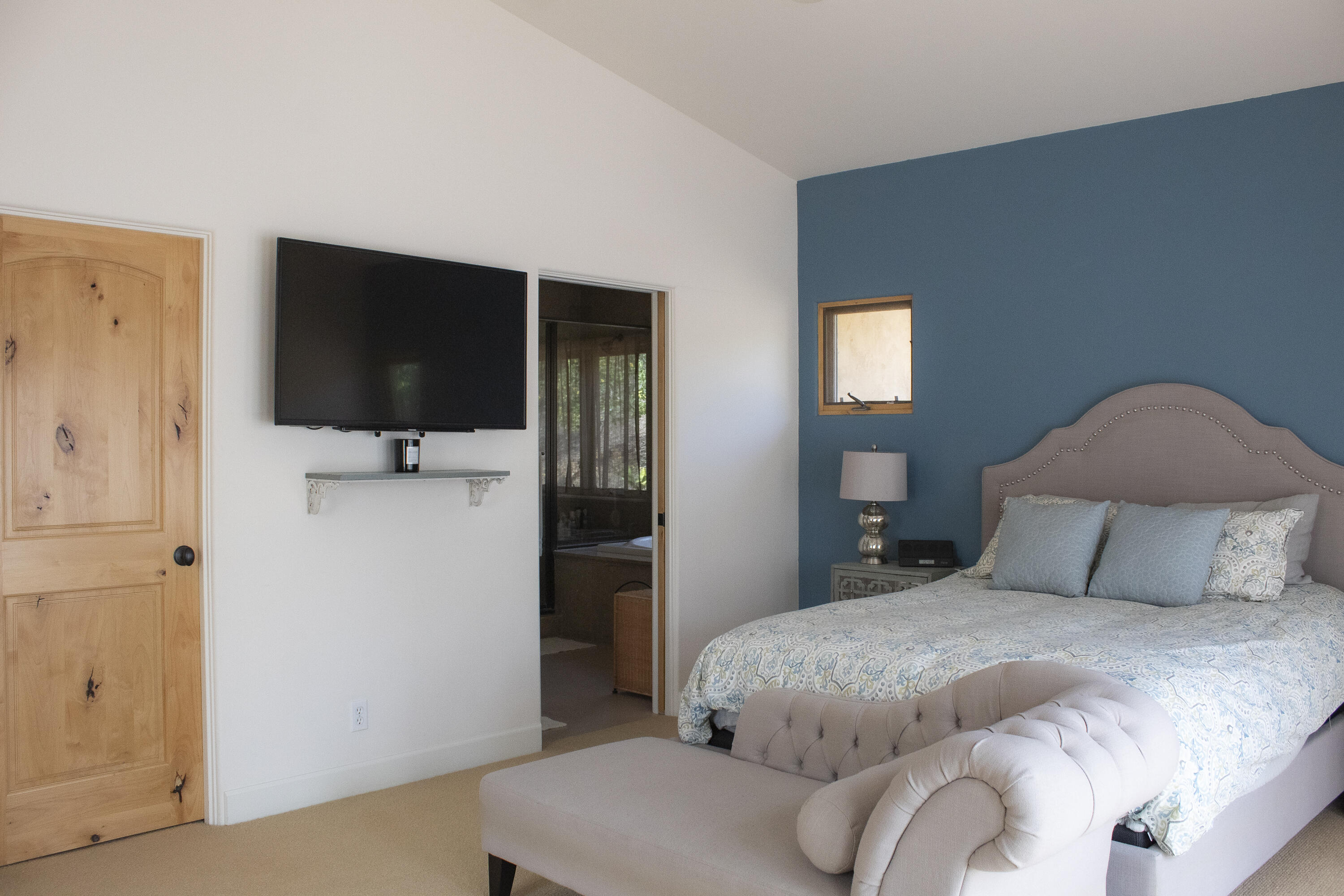Bedroom with a blue accent wall, patterned bedding, upholstered bench, and wall-mounted television.