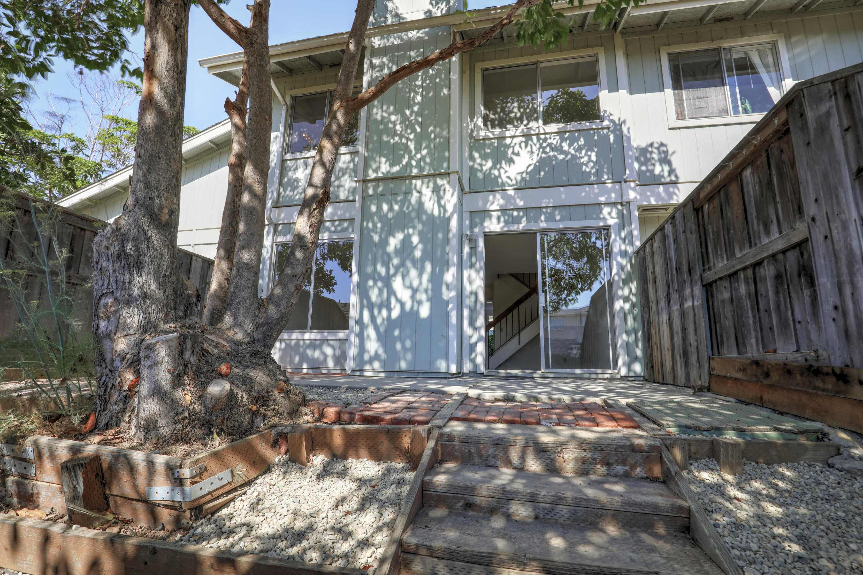 Two-story home exterior with a shaded patio, mature tree, and wooden walkway to a sliding glass door.