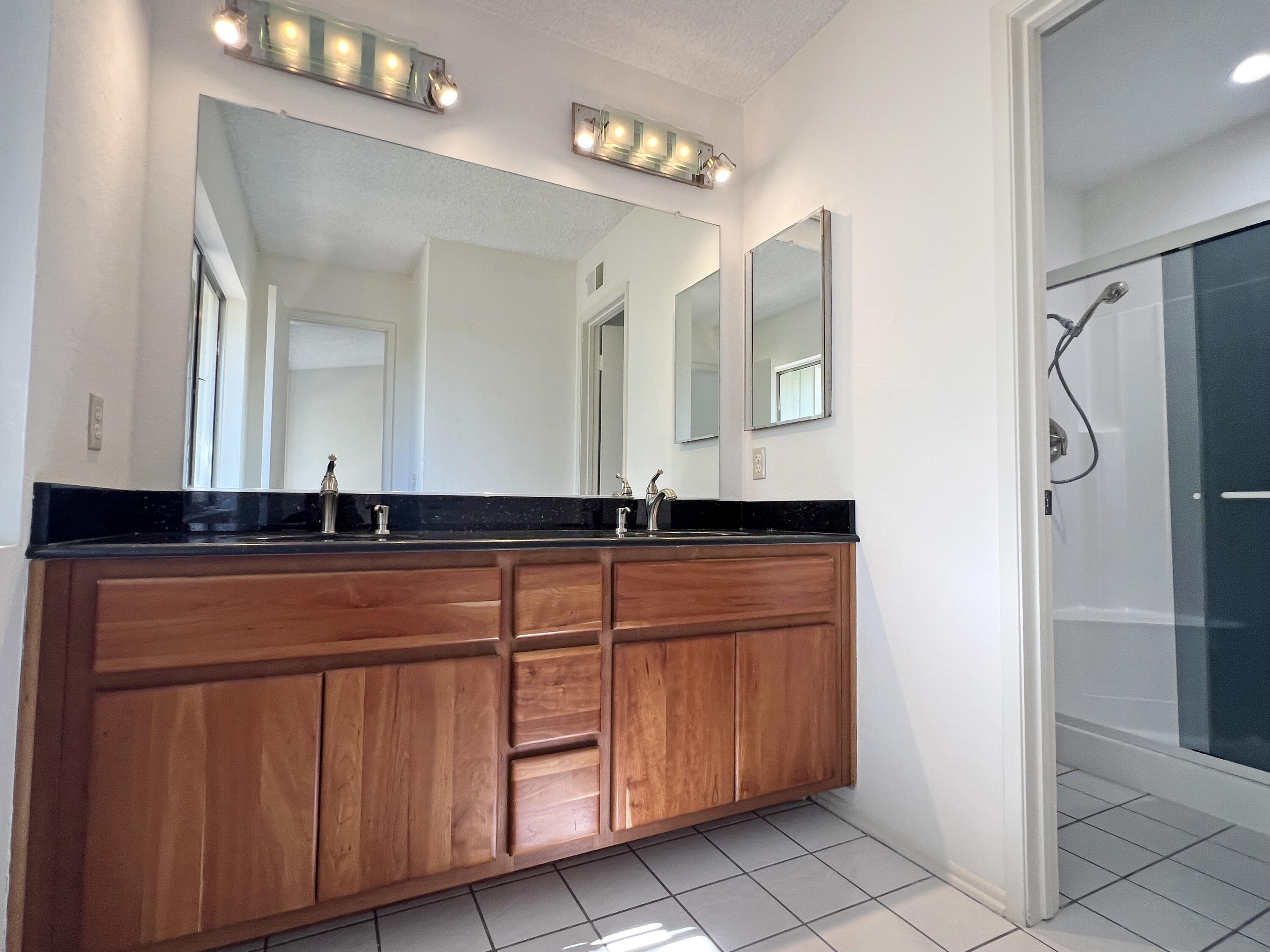 Bathroom with a wood double-sink vanity and black countertop beside a glass-enclosed shower.