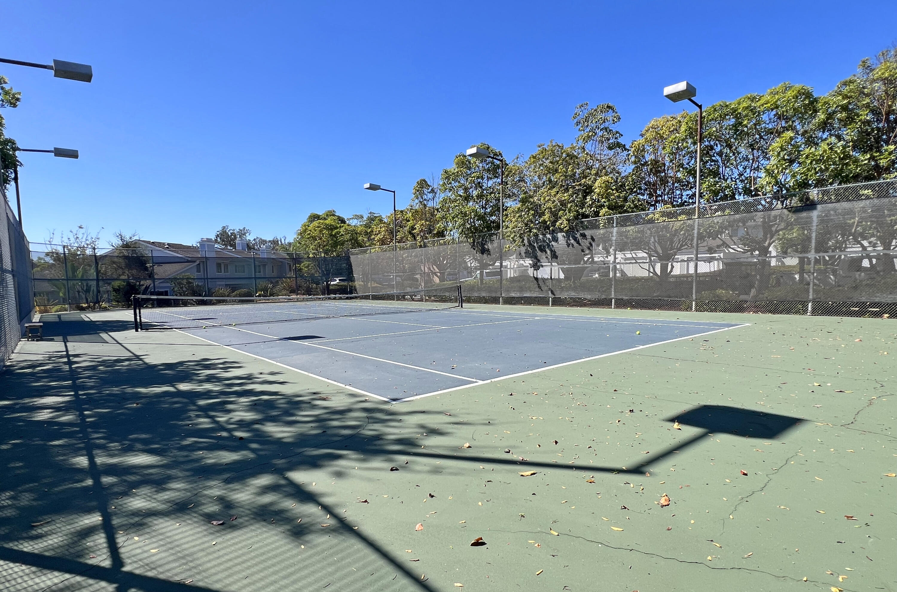Outdoor tennis court with tall fencing, overhead lights, and scattered leaves on the playing surface.