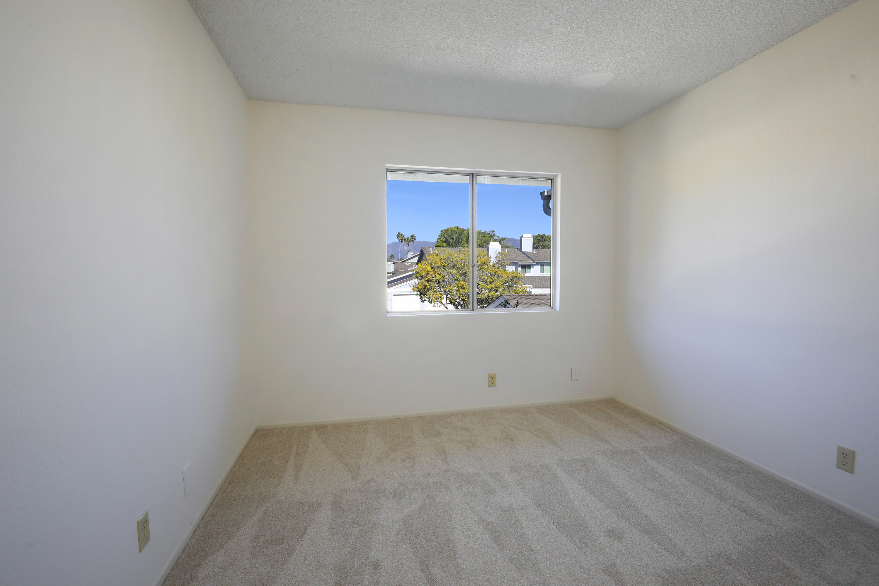 Empty carpeted bedroom with a window overlooking rooftops, trees, and distant mountains.