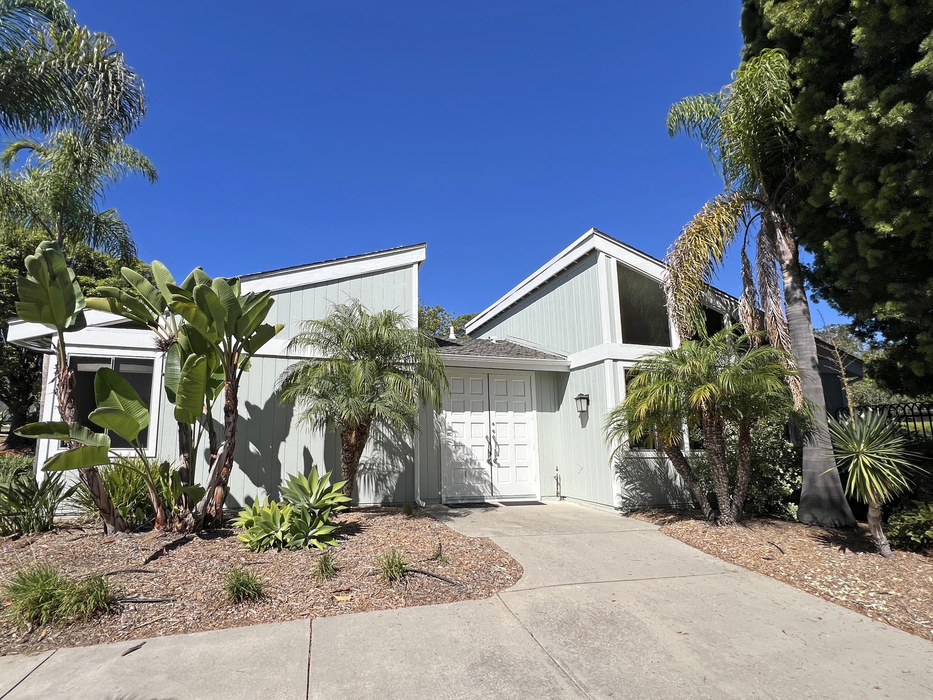 Light-colored two-story home with white double doors, a concrete walkway, and tropical landscaping.