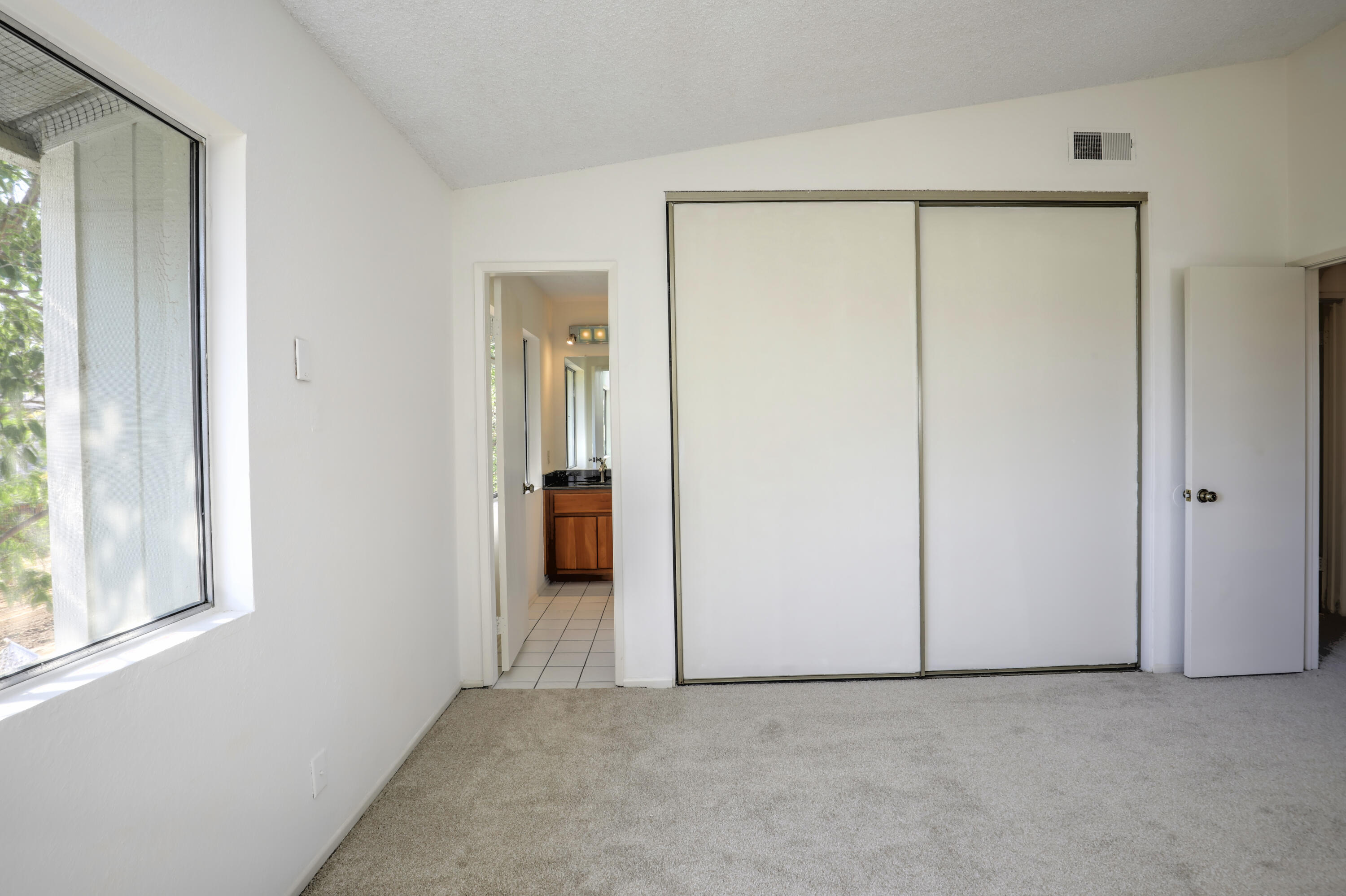 Vacant bedroom with beige carpet, sliding closet doors, and a window beside the bathroom doorway.