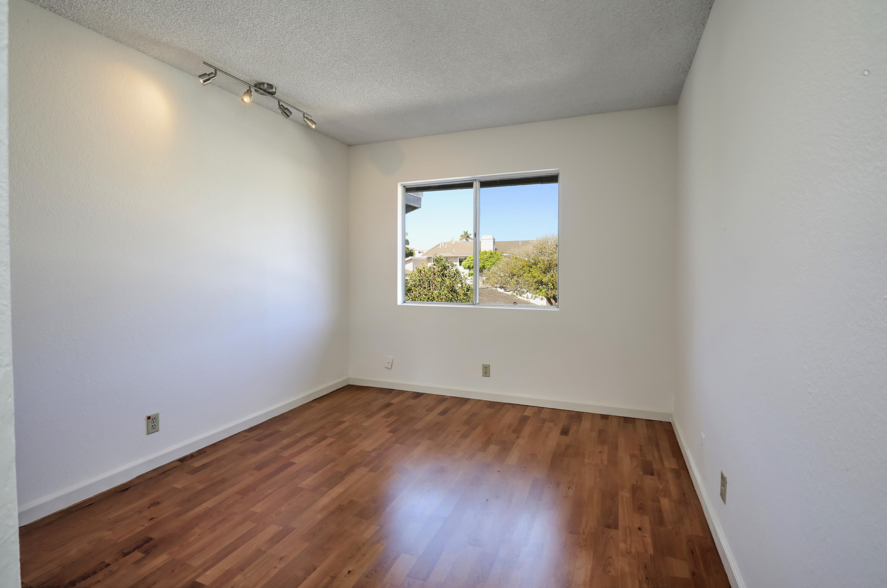 Empty room with hardwood floors, ceiling track lights, and a window overlooking trees and rooftops.