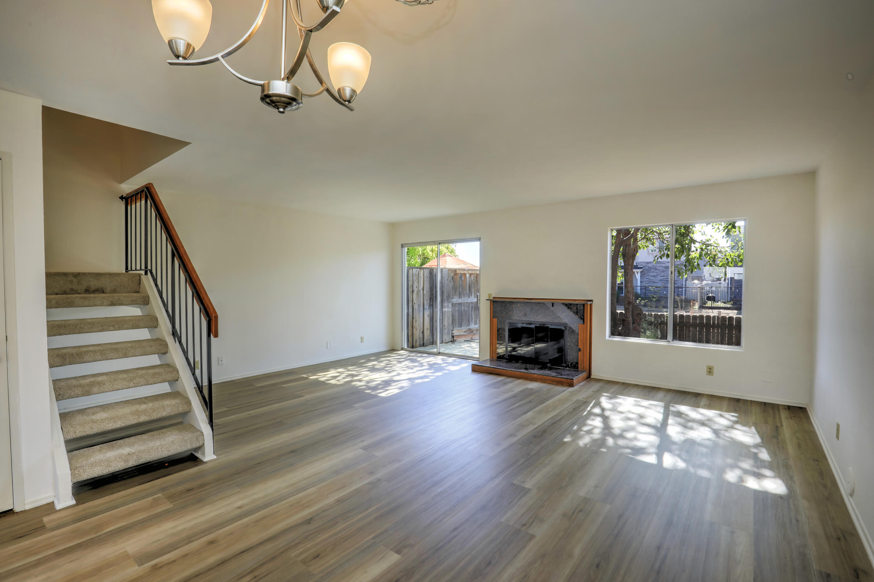 Empty living room with a fireplace, carpeted staircase, and sunlight streaming across wood-look floors.