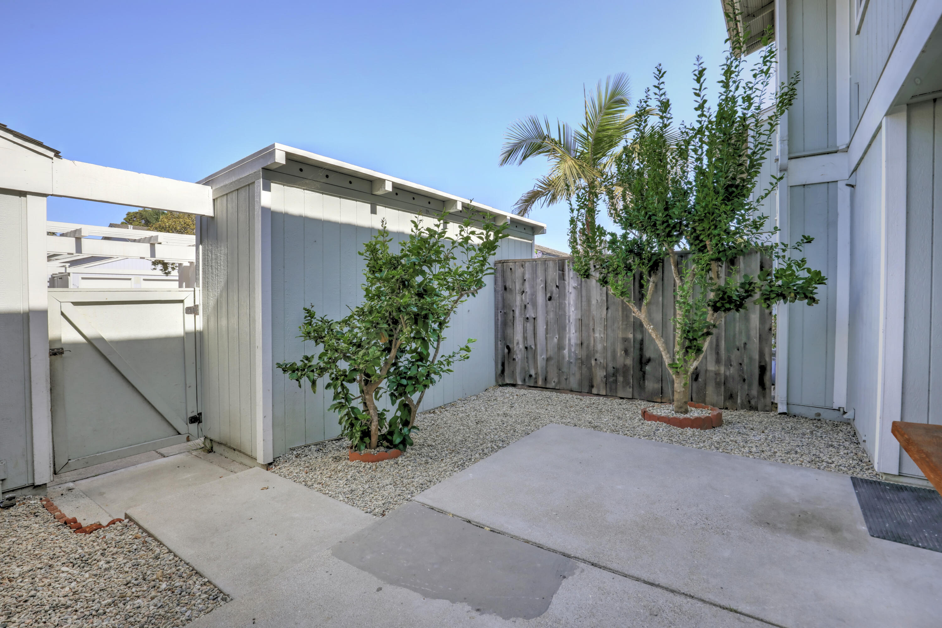 Enclosed courtyard with a concrete patio, gravel landscaping, leafy trees, and white fencing.