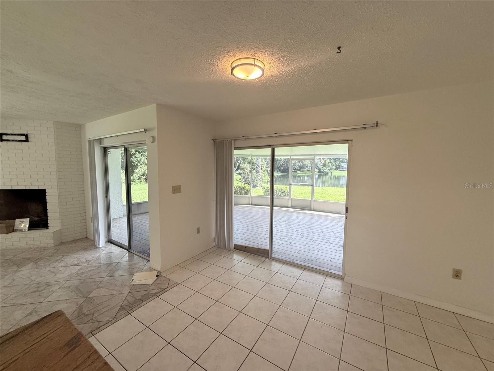 Vacant tiled living room with a white brick fireplace and screened patio overlooking a pond.
