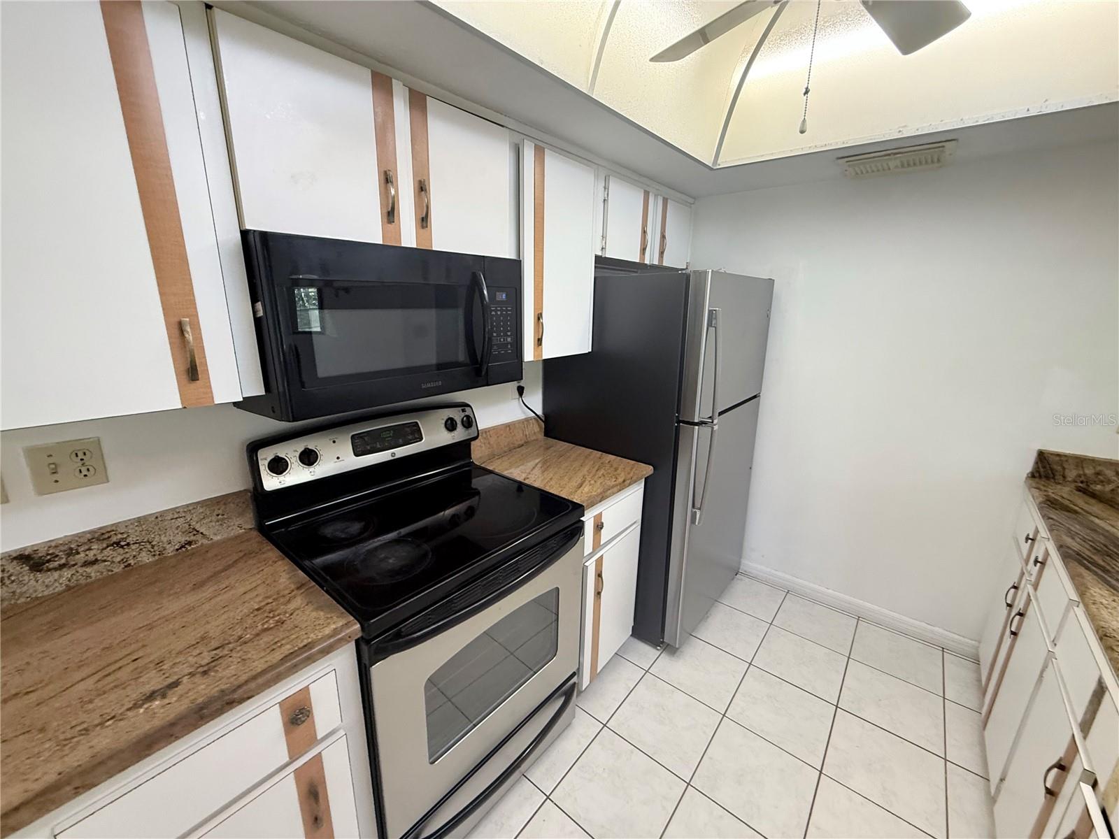 Galley kitchen with white cabinets, brown countertops, black range and microwave, and stainless-steel refrigerator.